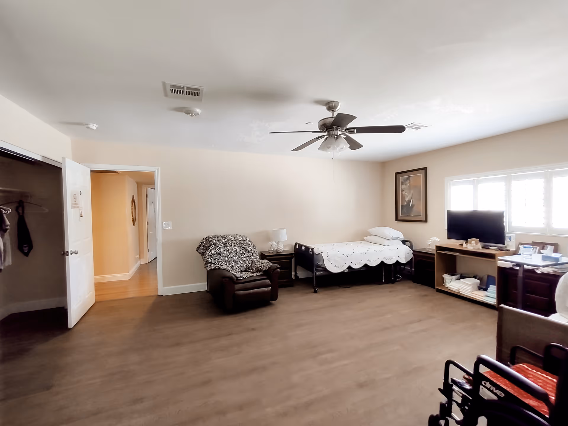 A spacious senior living bedroom with a bed, armchair, TV stand, and wheelchair nearby.