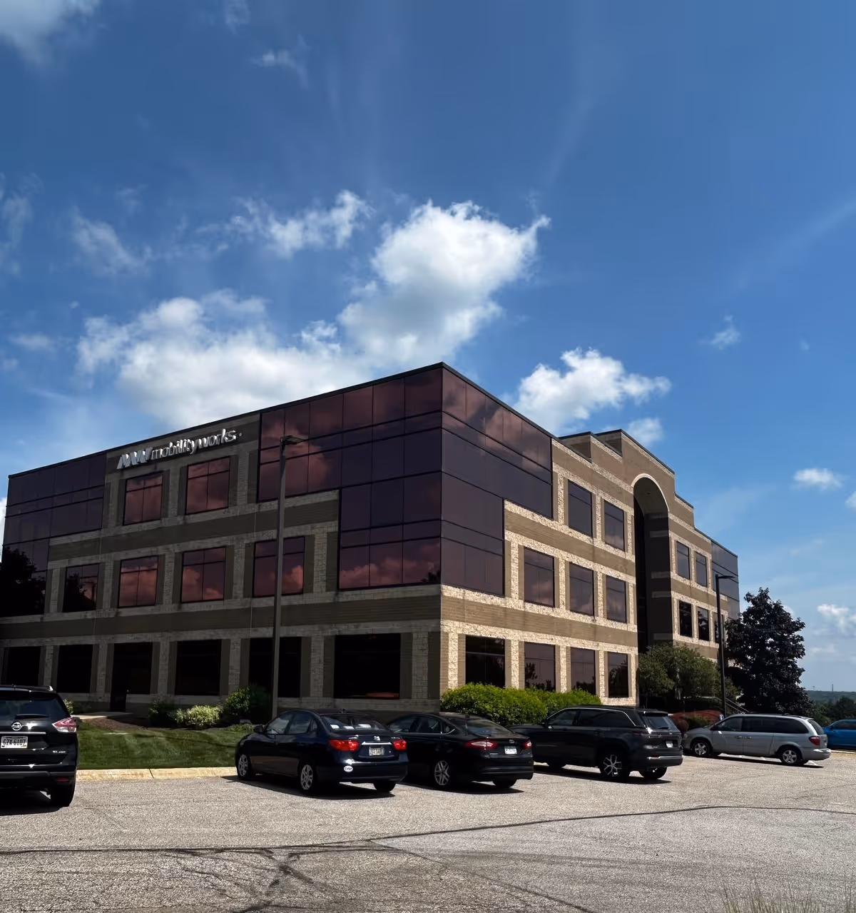 Three-story office building with glass panels, brick accents, and a parking lot under a blue sky.