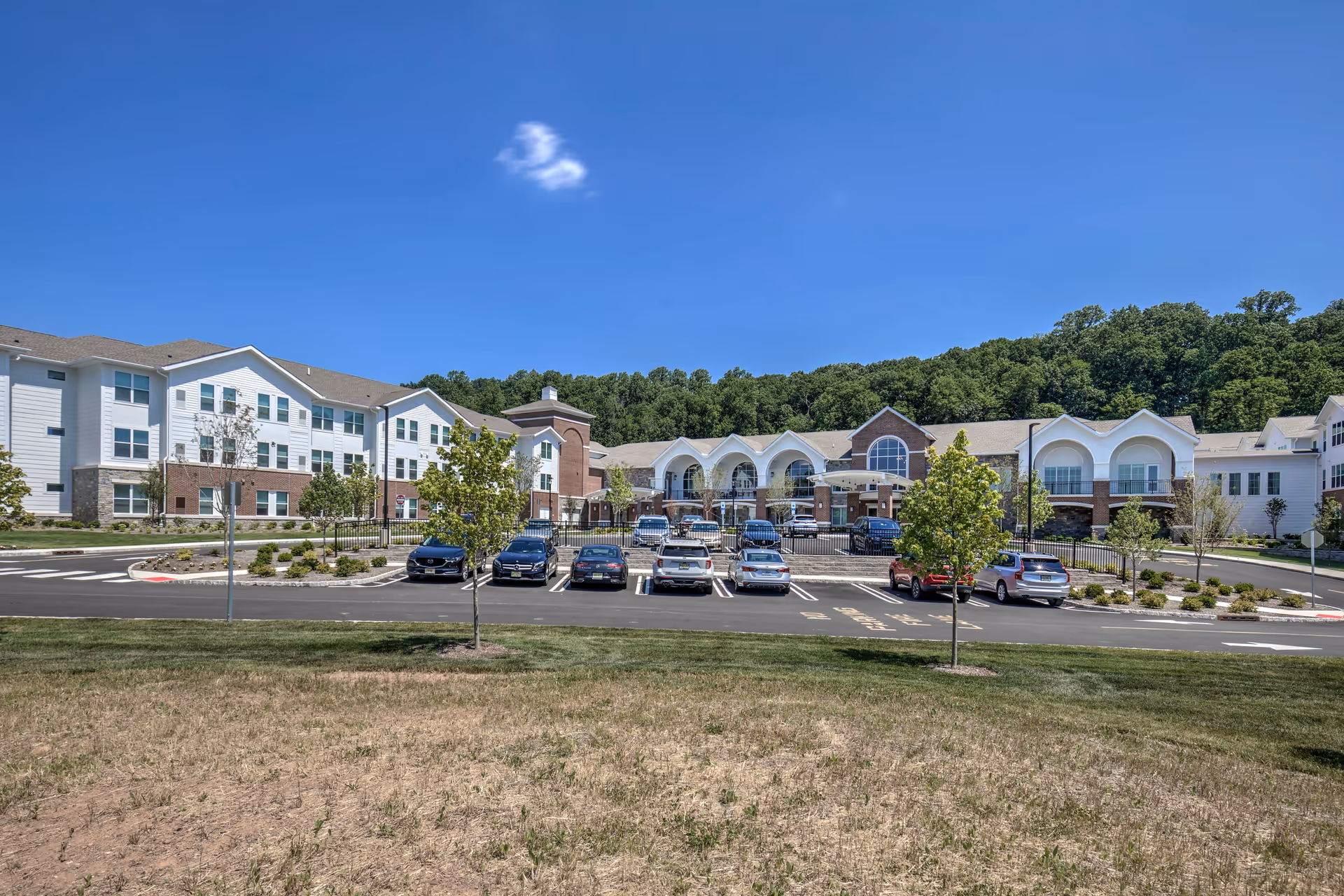 Exterior view of The Delaney of Bridgewater senior living facility showing a large, multi-story building with white and brick facade, several parked cars in front, small trees, and a clear blue sky with a single small cloud.
