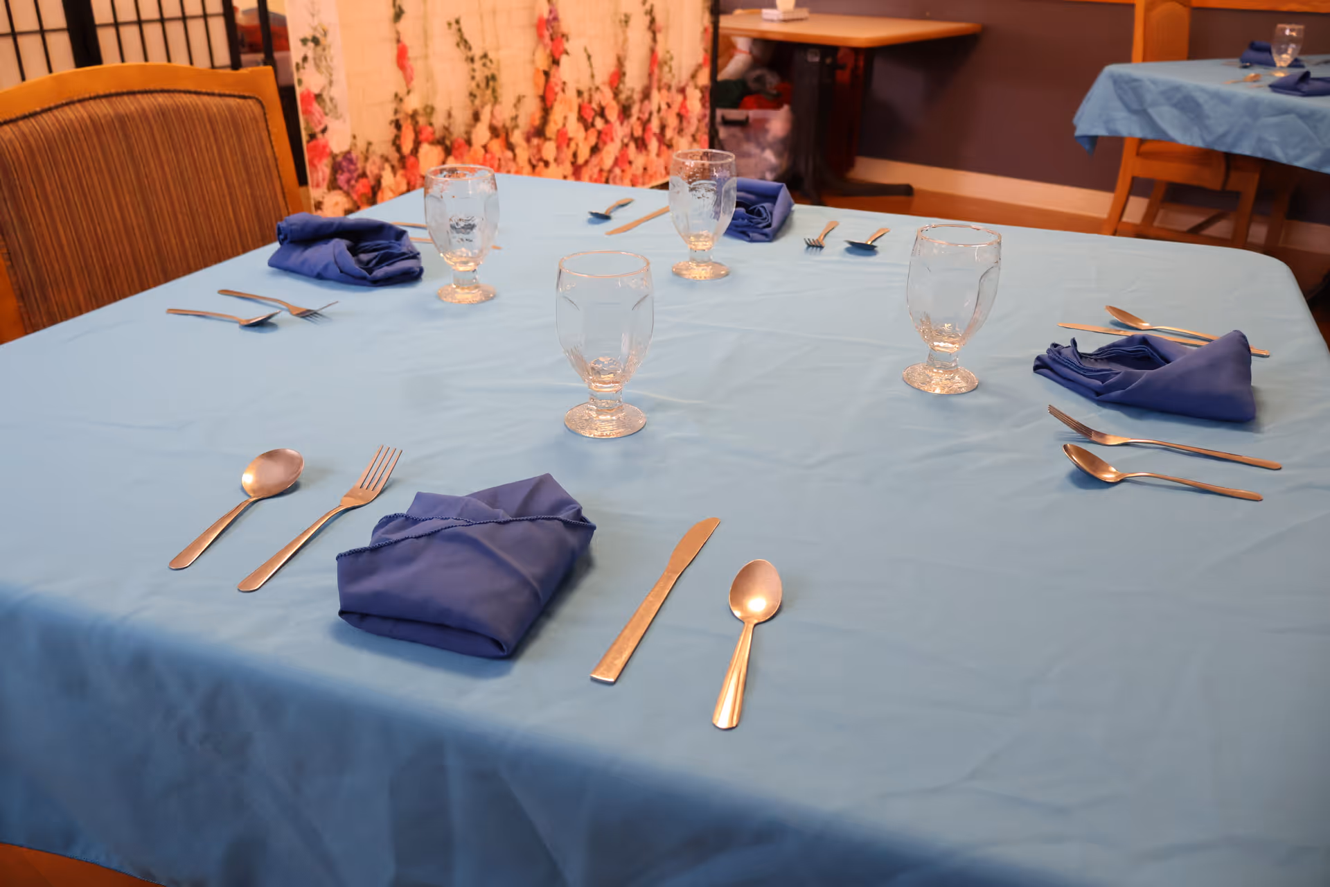 A dining table set with a light blue tablecloth, four clear water glasses, blue cloth napkins, and silverware including forks, knives, and spoons arranged neatly around the table. Chairs are positioned around the table and a floral curtain is visible in the background.