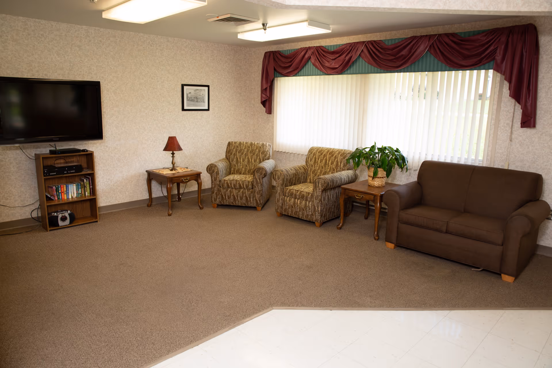 A small lounge with two patterned armchairs, a brown loveseat, side tables, a TV on a stand, and a large window with vertical blinds and a maroon valance.
