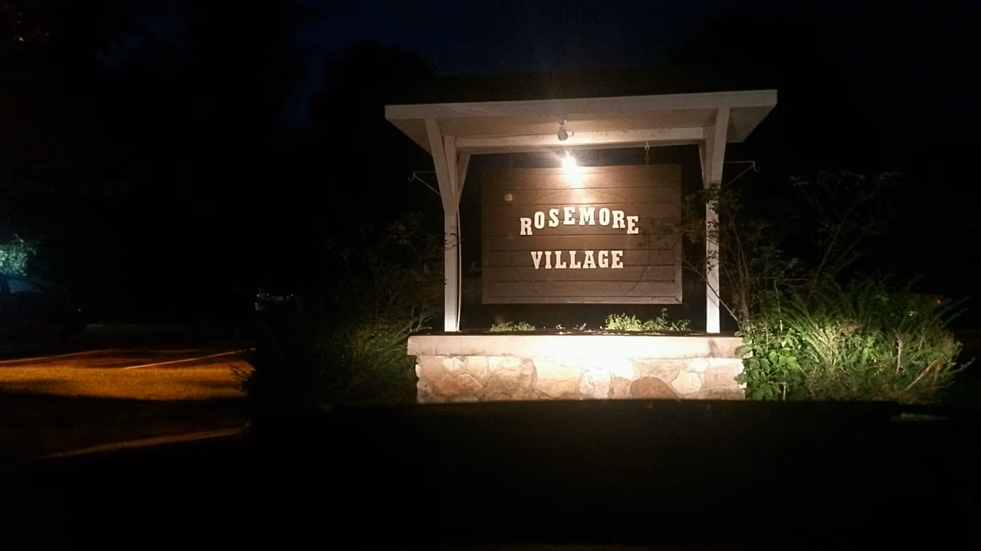 Nighttime exterior sign for Rosemore Village with stone base and surrounding shrubs.