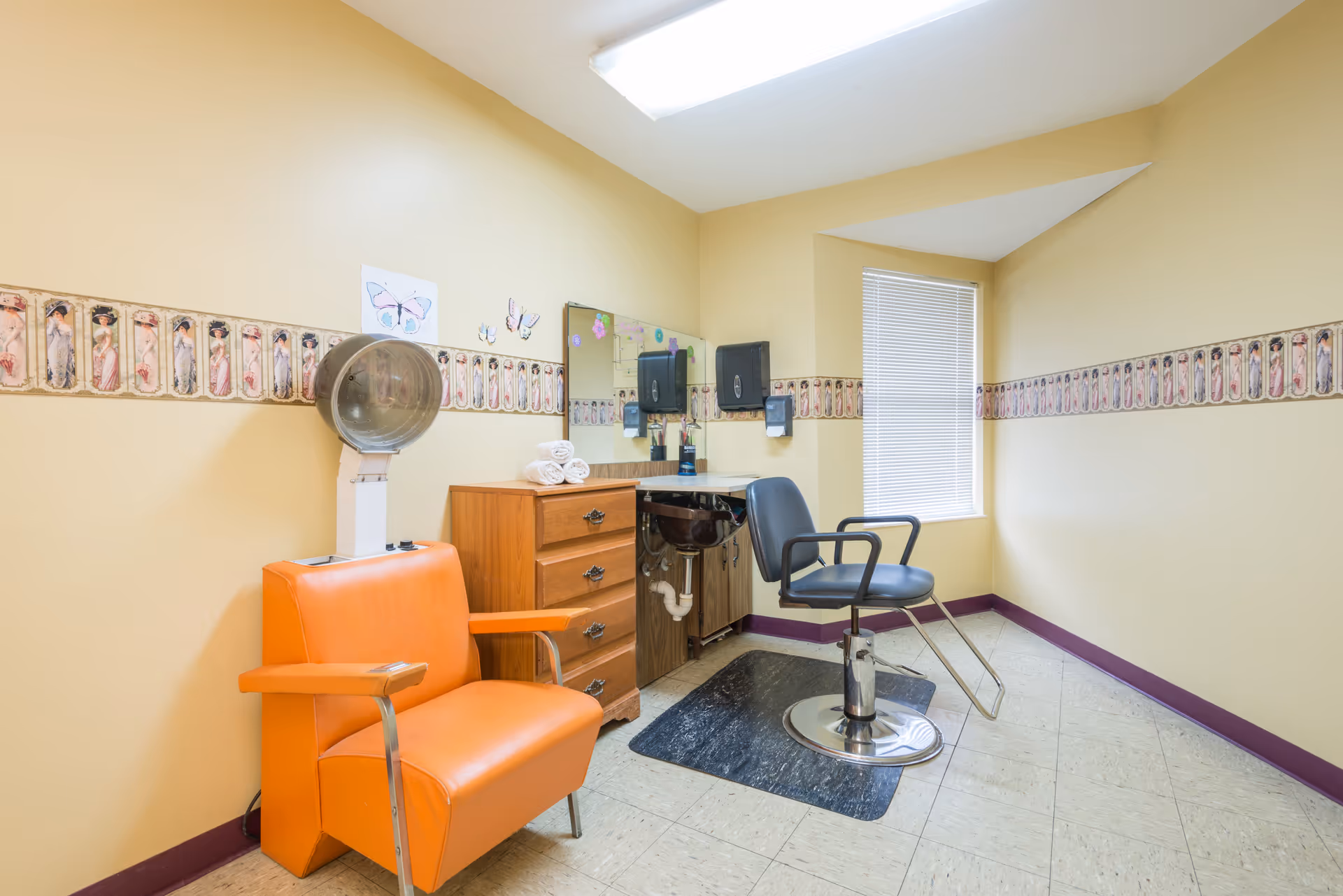 A small, yellow-walled grooming room with orange and black chairs, mirror, and sink.