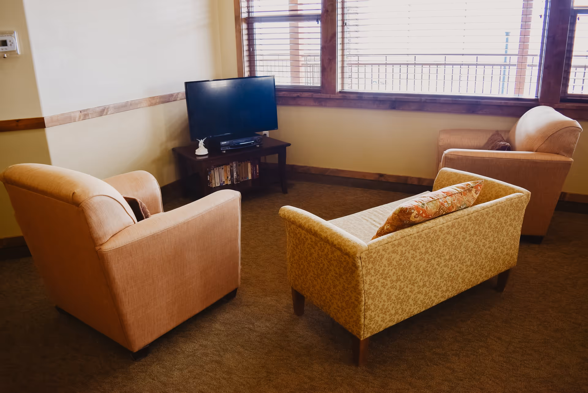 A cozy living room area with two beige armchairs and a patterned loveseat arranged around a small wooden TV stand holding a flat-screen television and some books. Large windows with wooden blinds let in natural light.