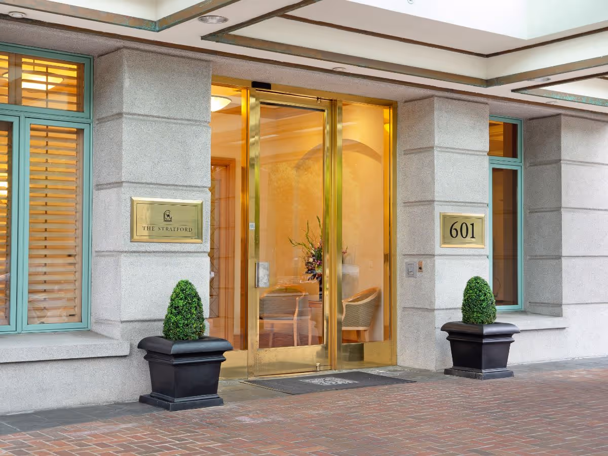 Entrance to The Stratford facility with golden glass doors, two potted topiary plants on either side, and a plaque displaying the name 'The Stratford' along with the number 601 on the wall.