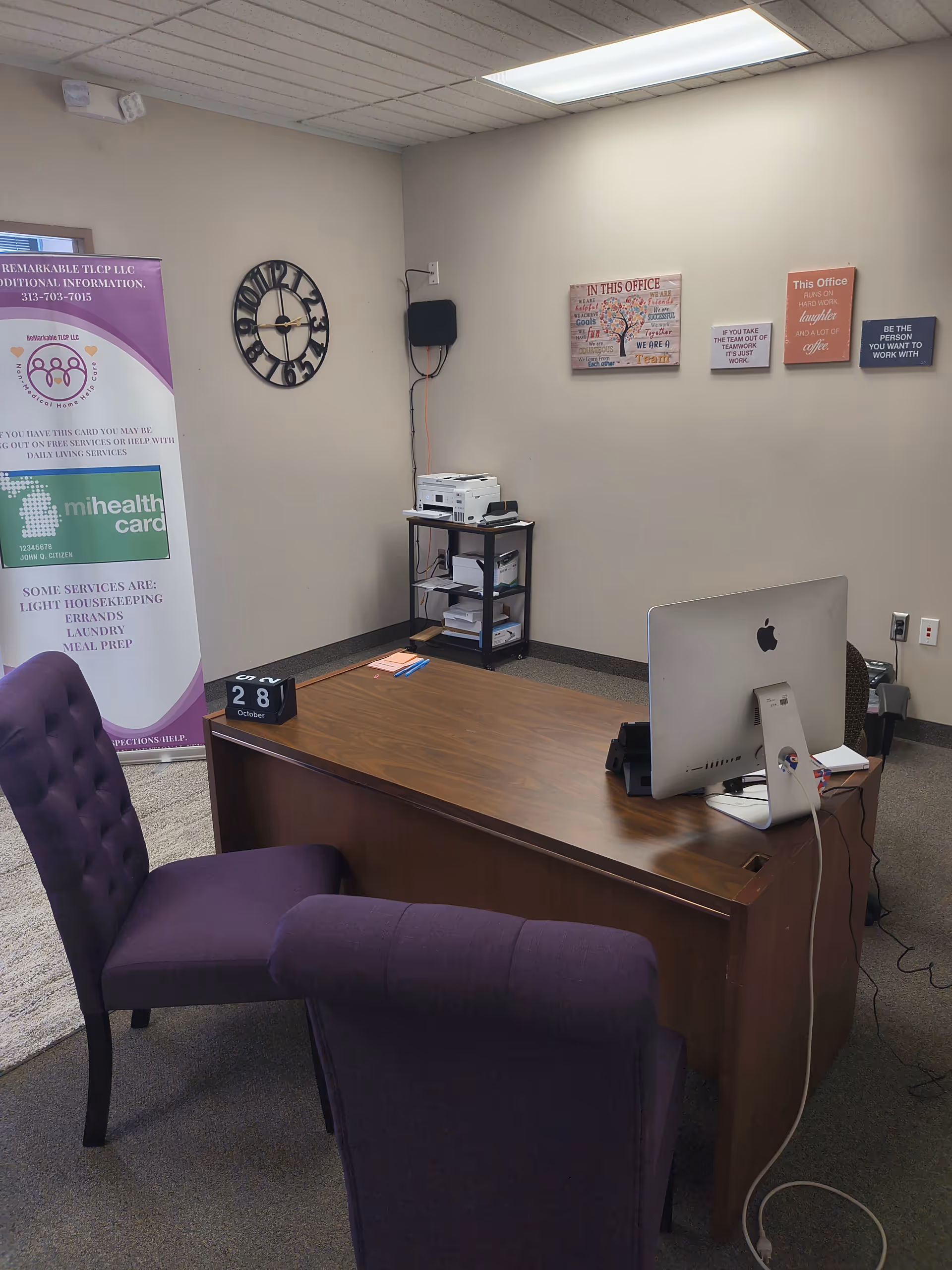 Office workspace with wooden desk, iMac, purple chairs, printer cart, and banners in a senior facility.
