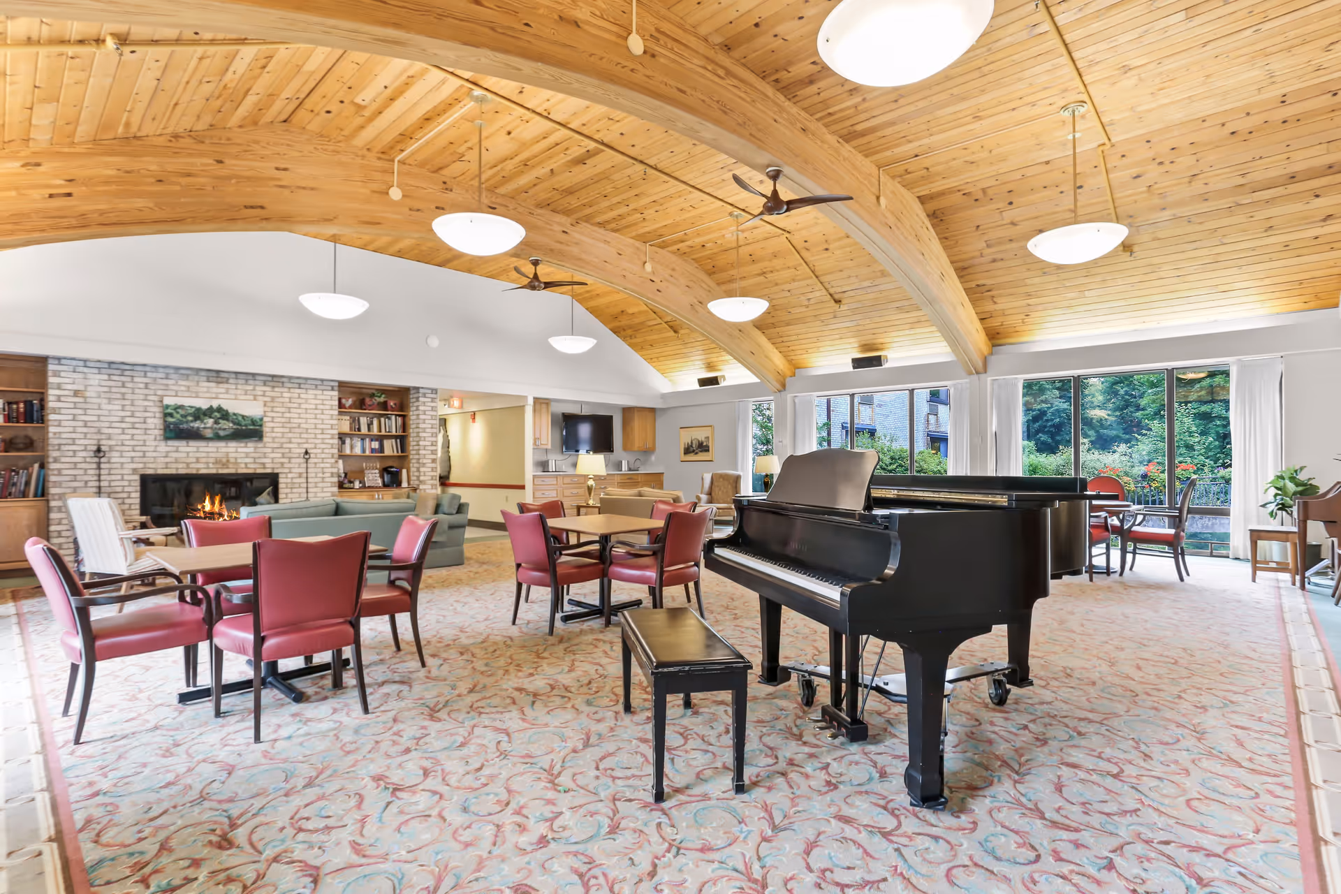 Spacious senior living common room with a grand piano, seating areas, fireplace, and vaulted wood ceiling.