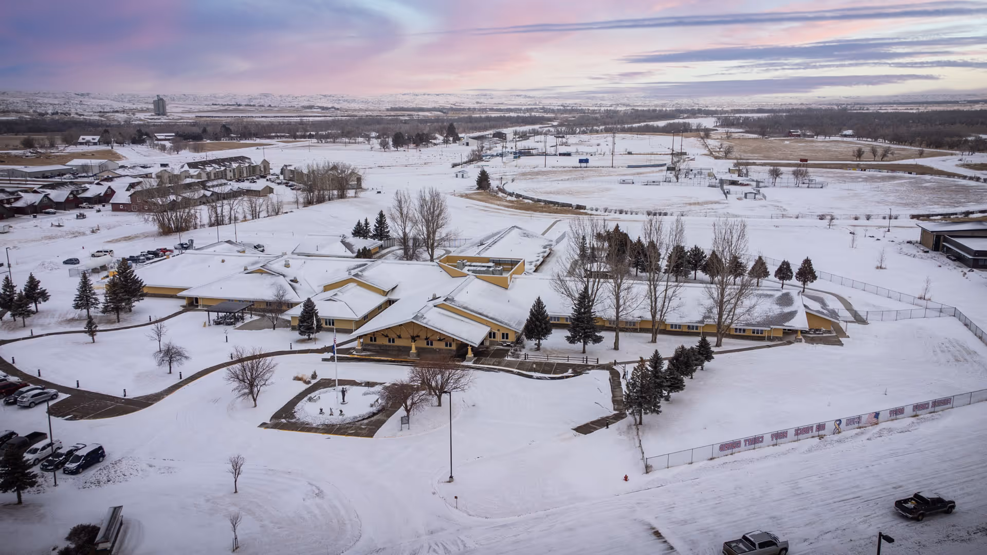 Aerial view of Eastern Montana Veterans Home covered in snow, showing a large single-story building with multiple wings surrounded by snow-covered grounds, trees, and parked cars. The landscape extends into the distance with a pastel-colored sky at sunset or sunrise.