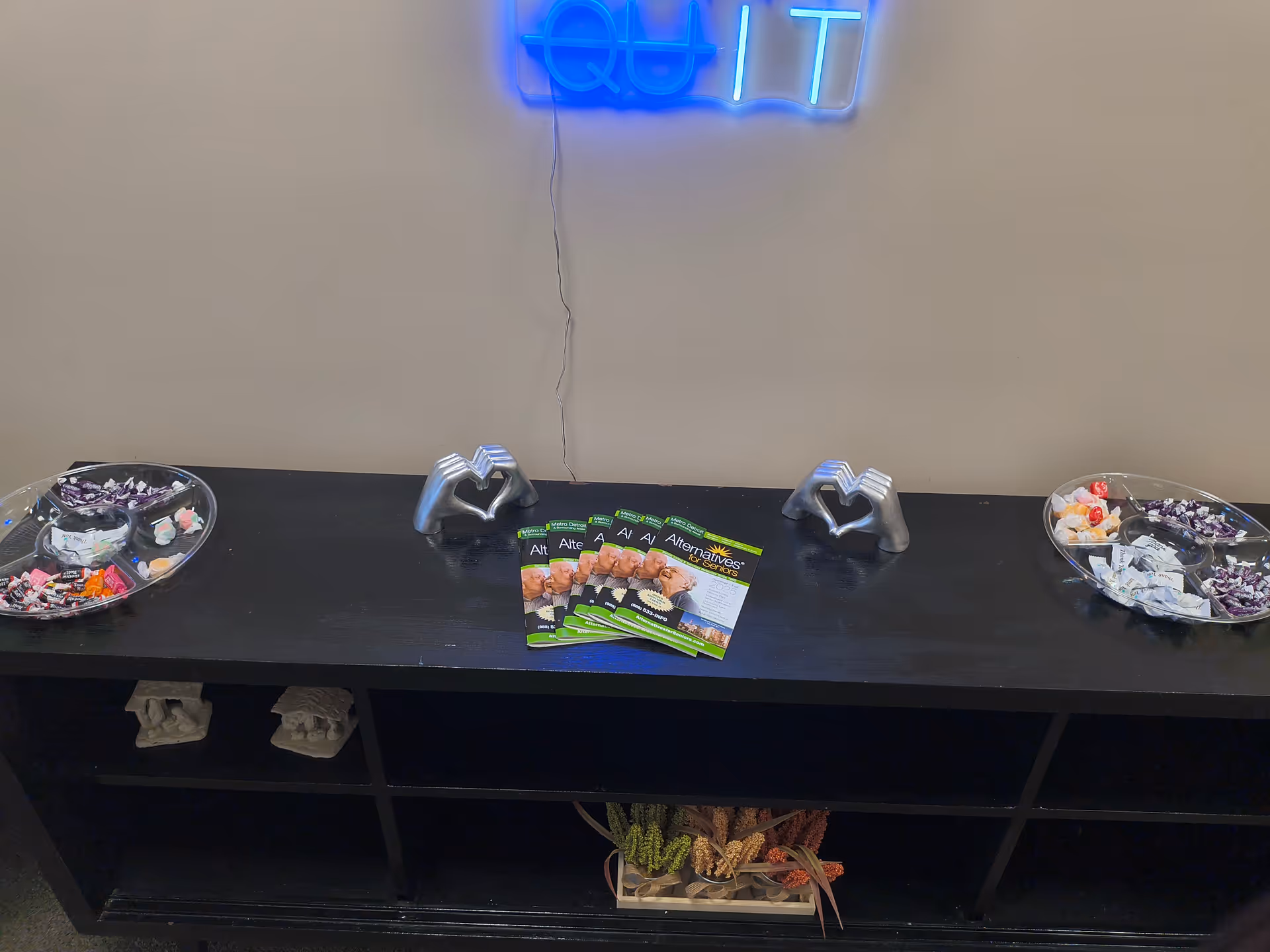 A black console table with two candy bowls, magazines, and a neon sign on the wall.