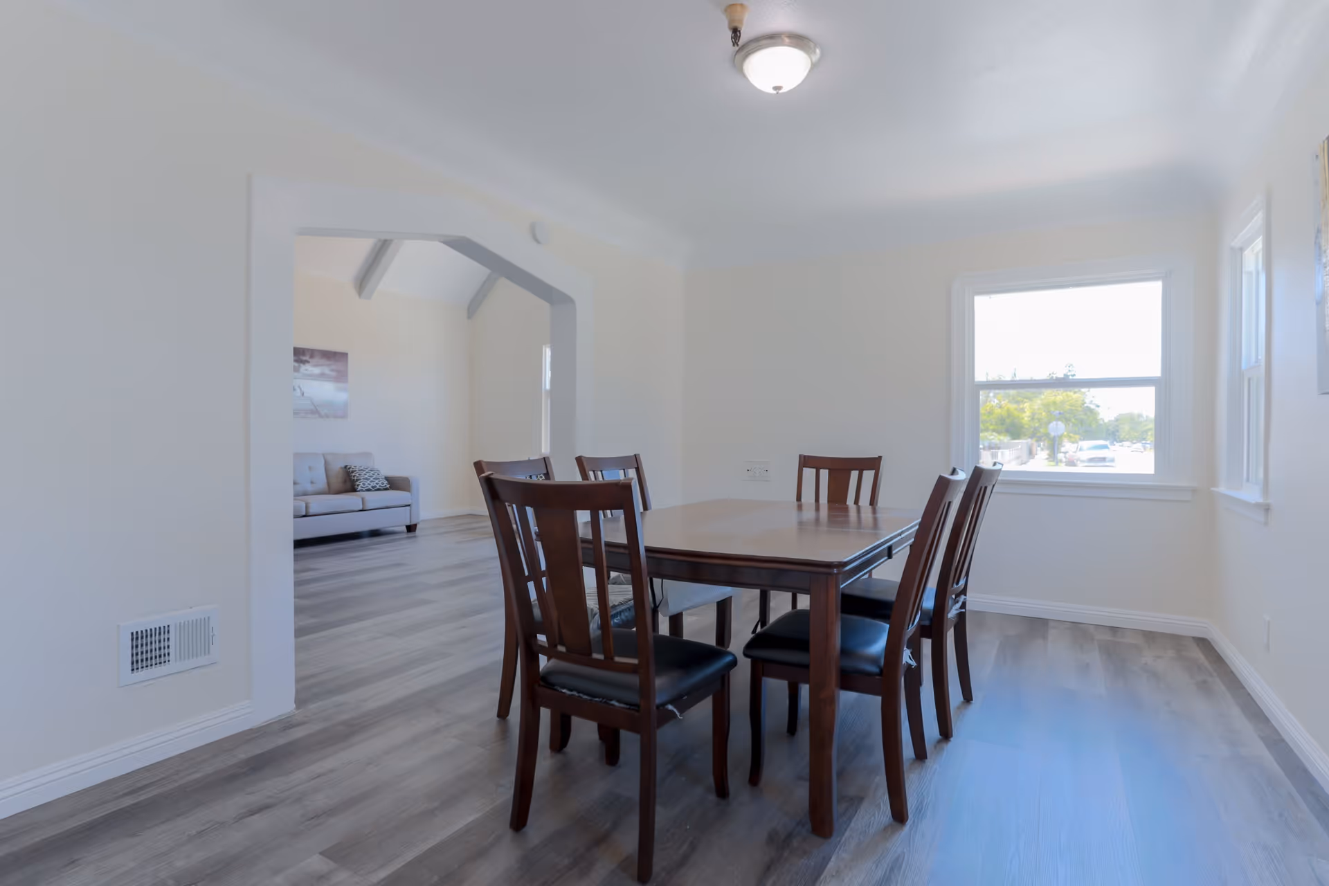A bright dining room with a wooden table and six matching chairs with black cushions. The room has light-colored walls and wood flooring. There is a large window letting in natural light and an open doorway leading to a living room area with a gray sofa and wall art.