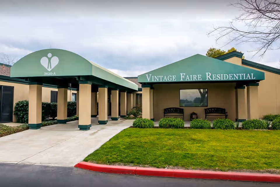 Front entrance of Vintage Faire Residential building with a green covered portico, benches, and landscaped lawn.