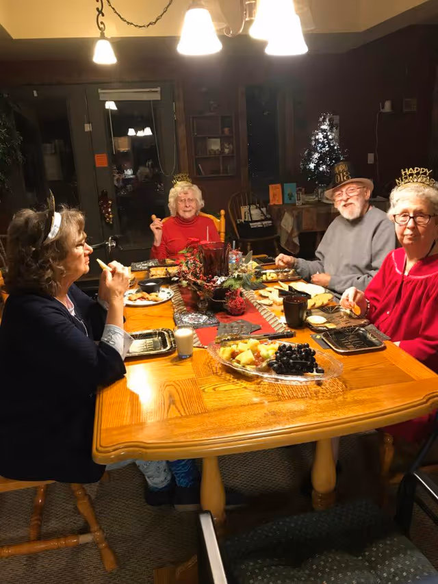 Four elderly people sitting around a wooden dining table enjoying a meal together. The table is set with plates of food including fruit and crackers, and there is a festive centerpiece. The individuals are wearing New Year's Eve hats and headbands, and the room is warmly lit with hanging lights above the table.