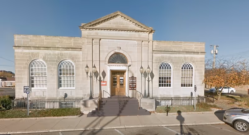 A stone building with arched windows and steps leading to a central entrance facing sidewalk.