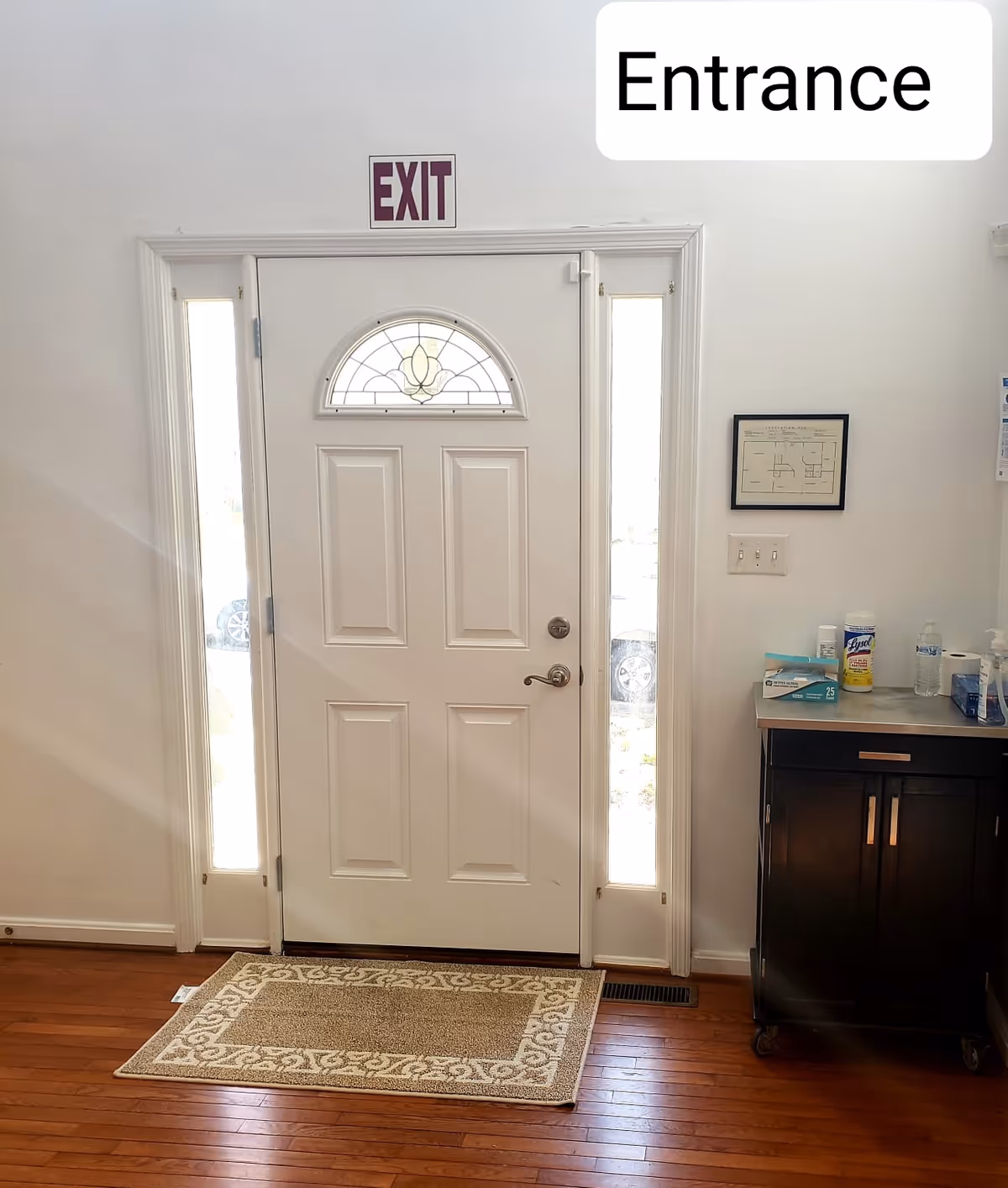 White interior entrance with exit sign above a paneled door, sidelights, and medical cart nearby.