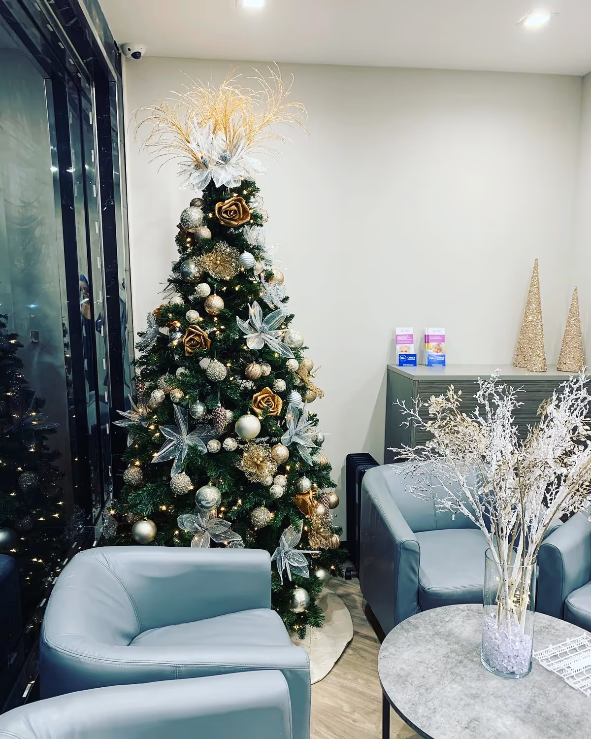 Festive Christmas tree with gold and silver ornaments beside blue lounge chairs in a modern interior.