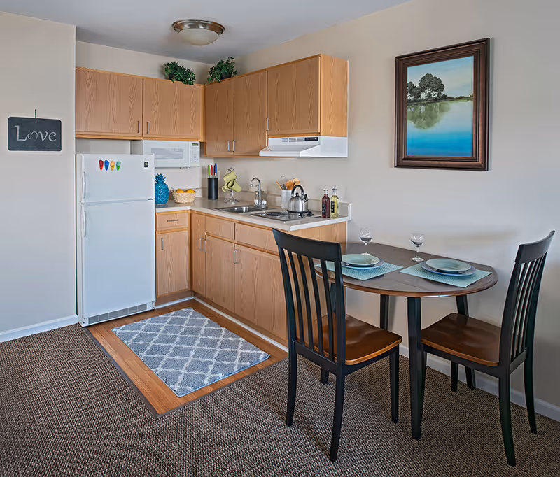 Compact kitchenette with wooden cabinets, appliances, and a small dining table for two.