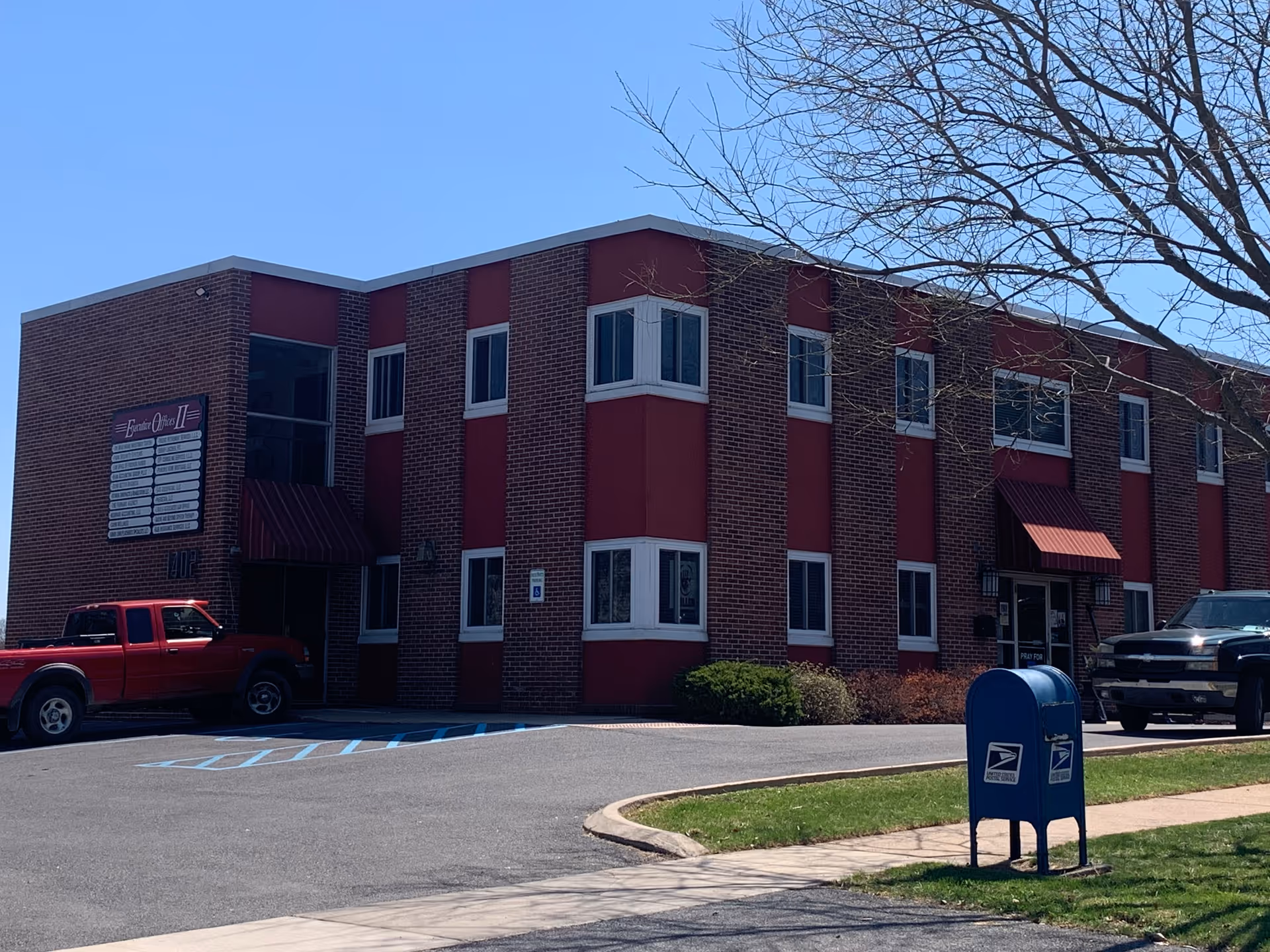 Two-story brick senior living facility with red accents, parked vehicles, and a blue mailbox.