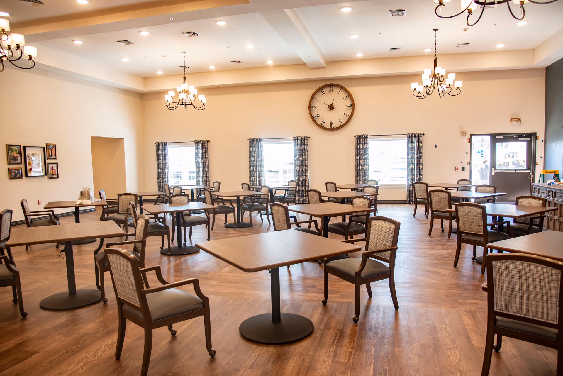 Spacious dining room with multiple tables, chairs, chandeliers, and a wall clock.