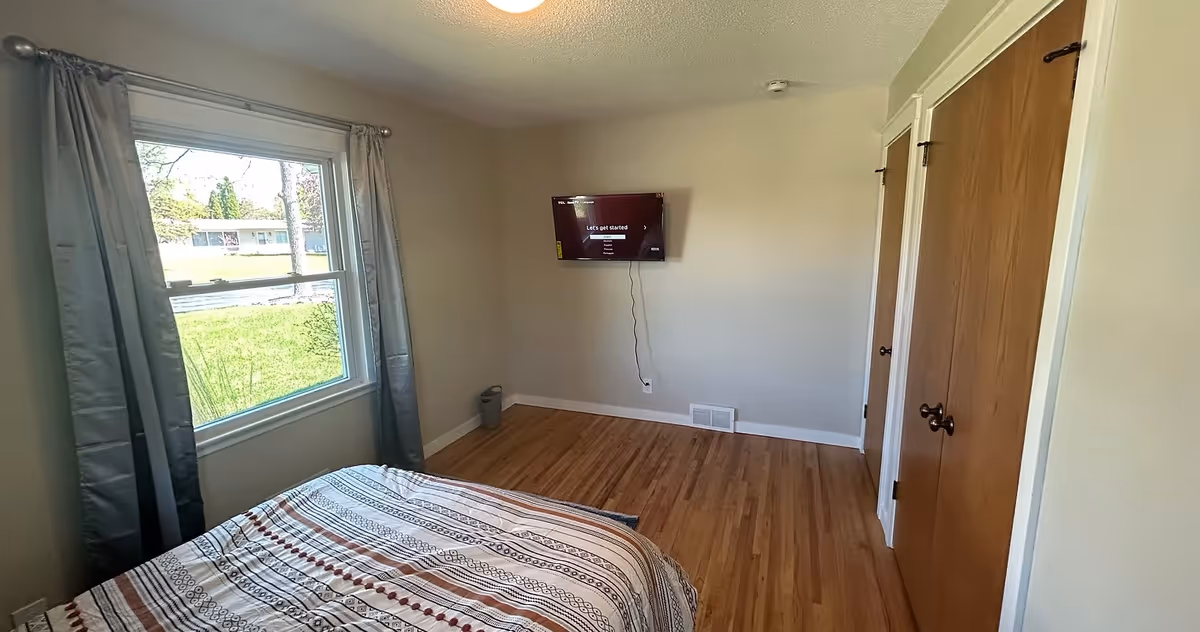 A cozy bedroom with a window, curtains, a bed, and a wall-mounted TV.
