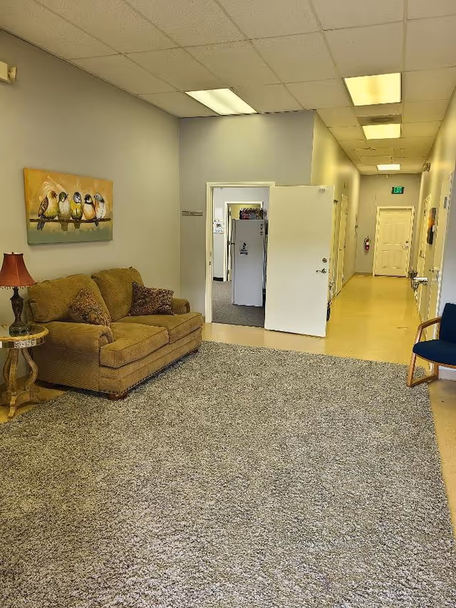 A cozy lounge area with a beige couch, lamp, and bird painting in a facility hallway.