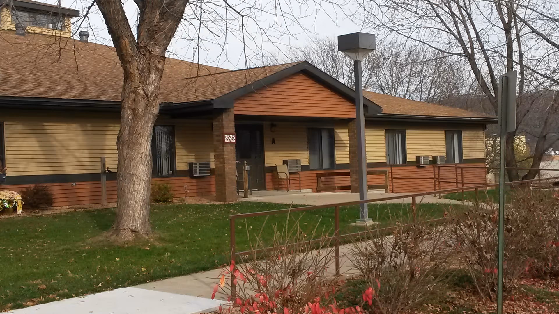 Exterior view of a single-story building with beige and brown siding, a sloped roof, and several windows with air conditioning units. There is a concrete walkway leading to the entrance, a large tree in the foreground, some bushes with red leaves, and a lamp post near the walkway. The building has a sign with the number 2525 and the letter A near the entrance.