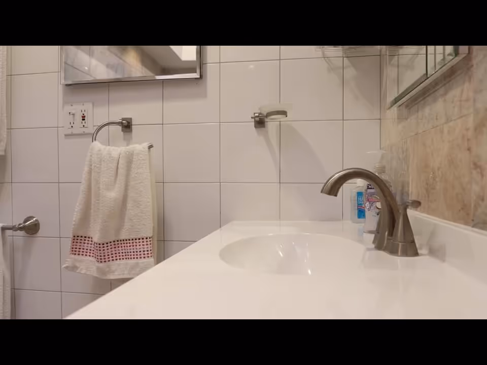 Bathroom sink with faucet, towel, mirror, and tiled walls in a senior living facility.