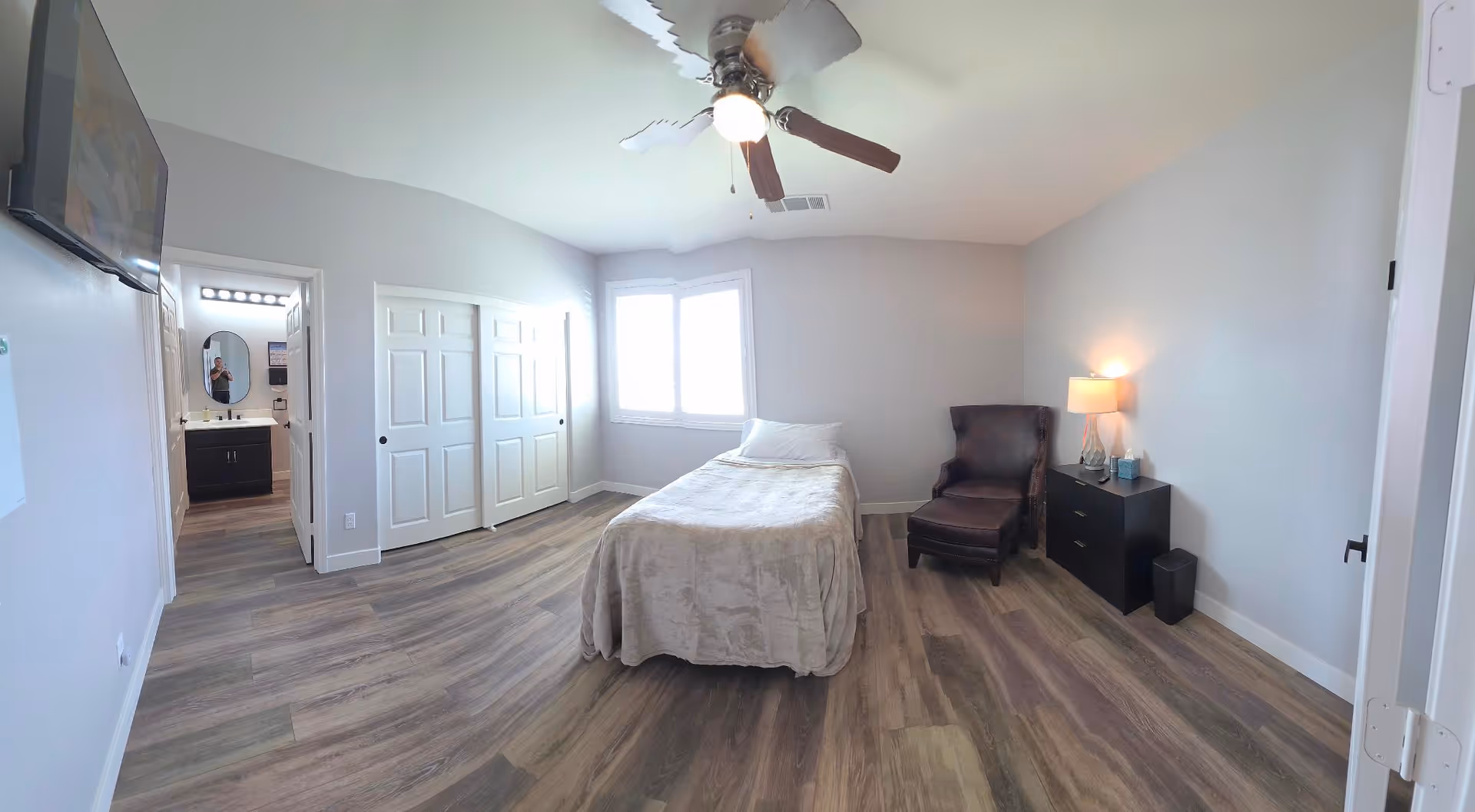 A light-gray bedroom with a single bed, window, ceiling fan, and dark furniture.