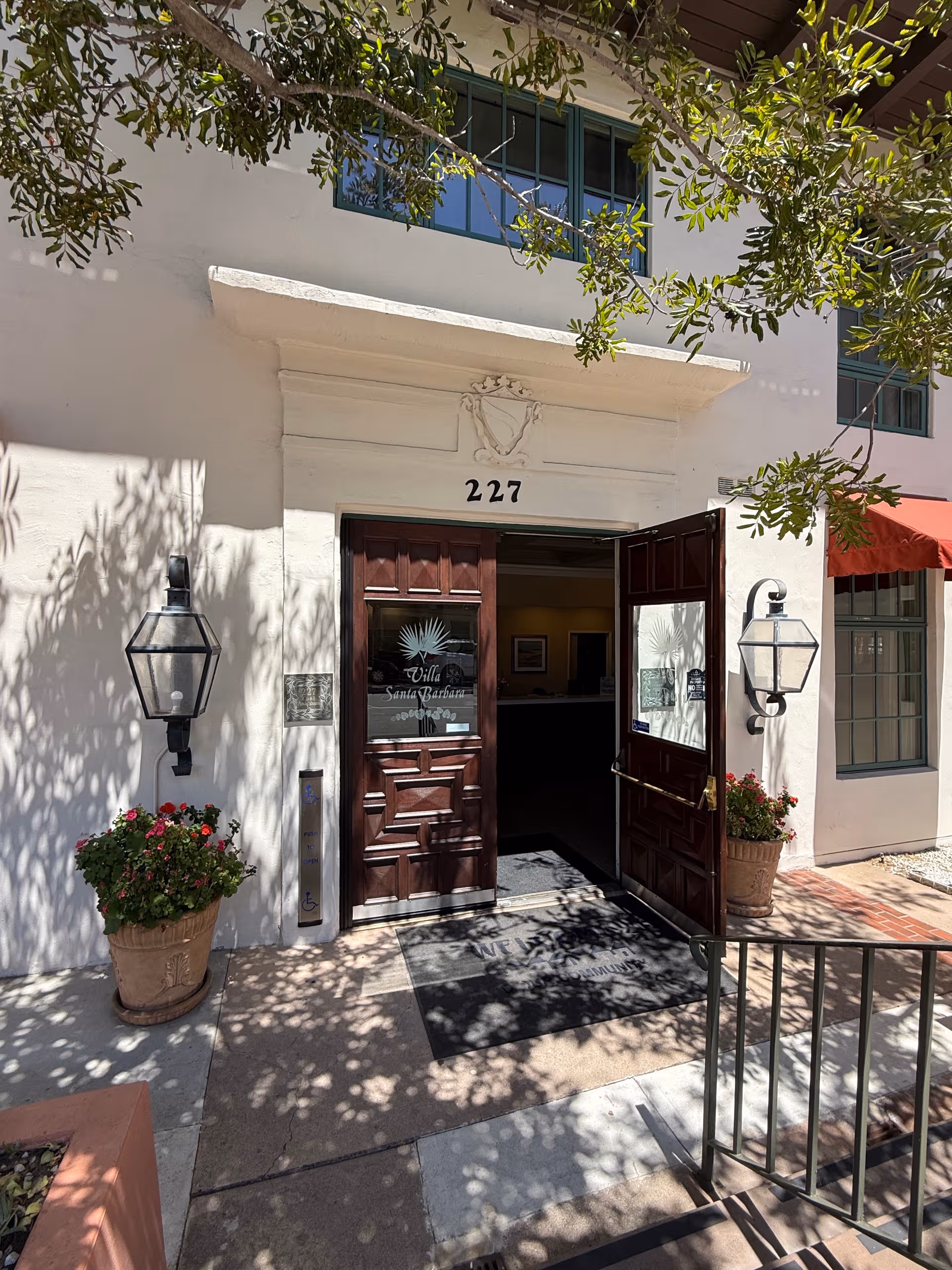 Villa Santa Barbara entrance with open wooden doors, lanterns, potted plants, and overhanging tree branches.