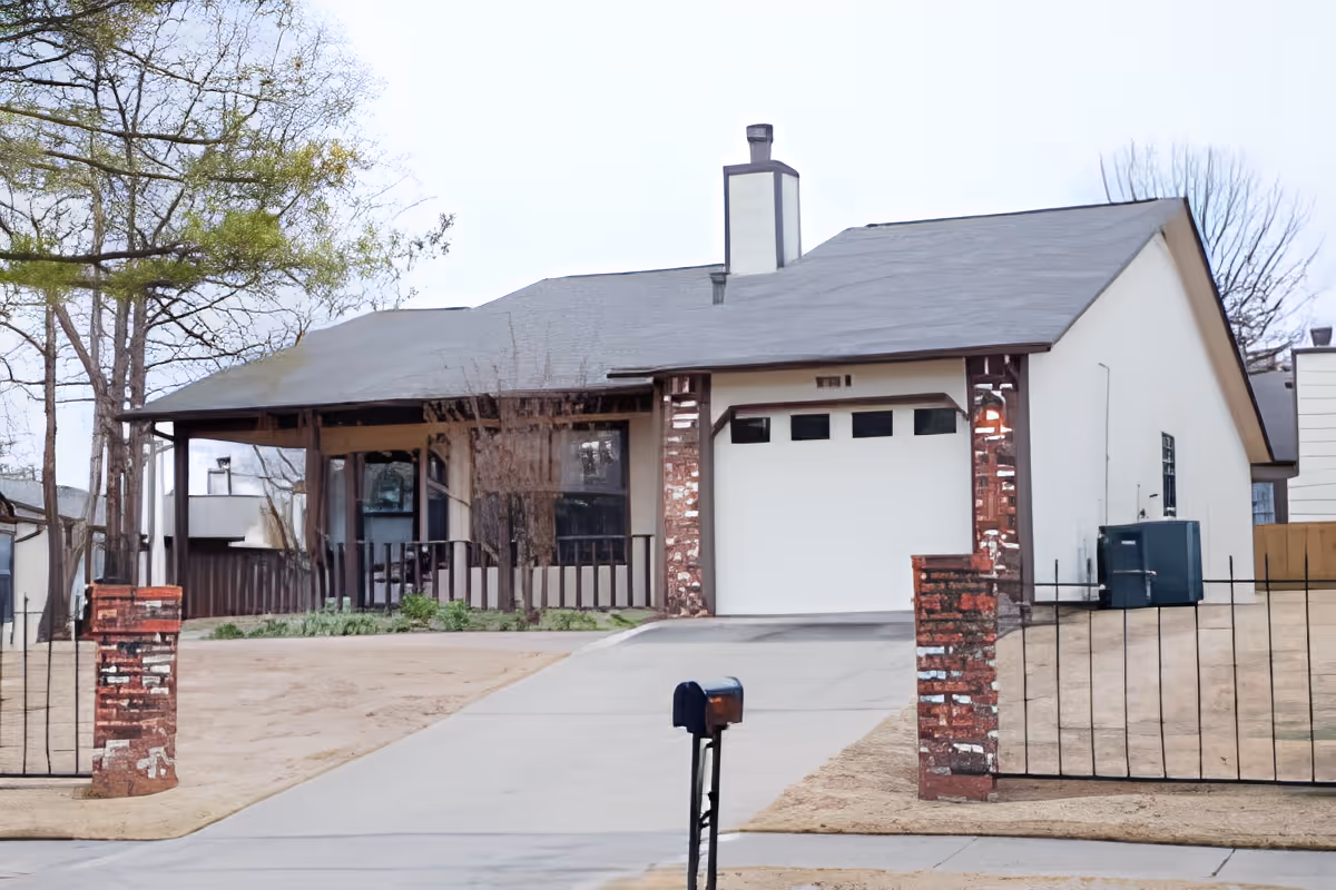Front exterior view of a single-story house with a sloped roof, a chimney, a garage with a white door, a concrete driveway, a black mailbox on a post, and a fenced yard with brick pillars. Trees and neighboring houses are visible in the background.
