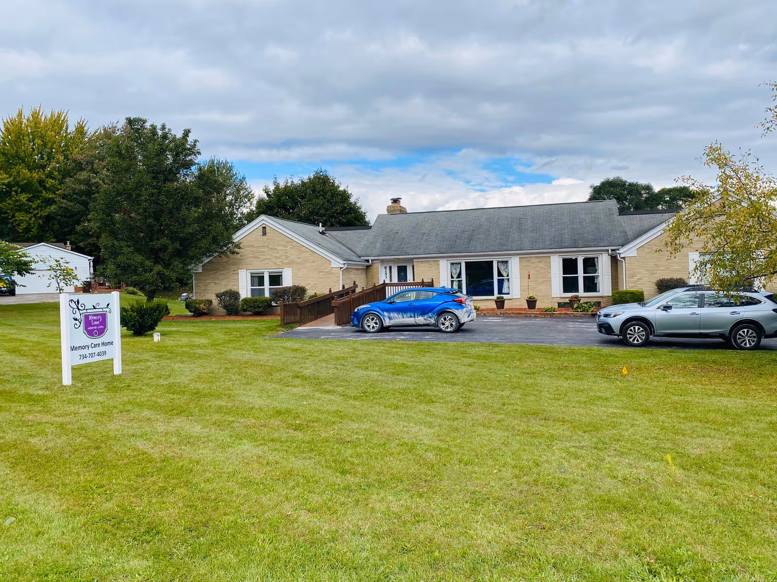 Front view of a single-story brick assisted living facility with a lawn, driveway, and two parked cars under a cloudy sky.
