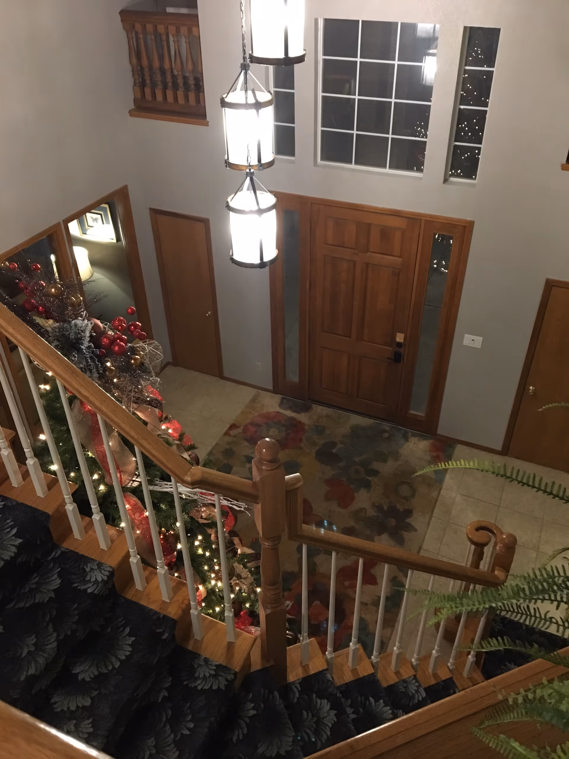 An upstairs view of a decorated foyer with a staircase, chandeliers, and holiday garlands.