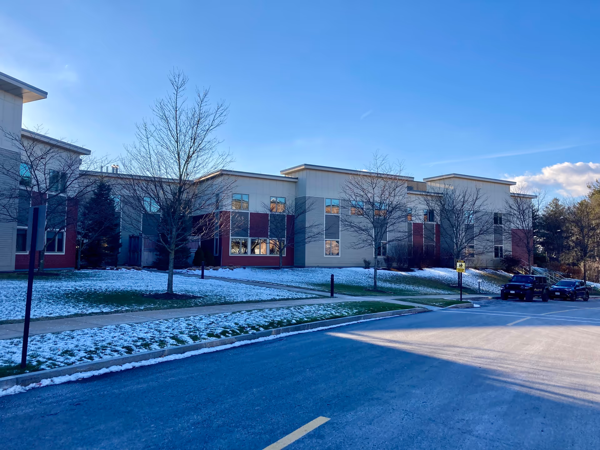 Two-story retirement community building exterior with bare trees, patches of snow on the lawn, and cars parked along the street under a clear blue sky.
