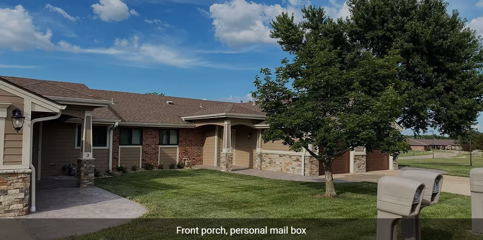 Single-story brick and siding senior living units with front porches, garages, a tree, and mailboxes on a sunny day.