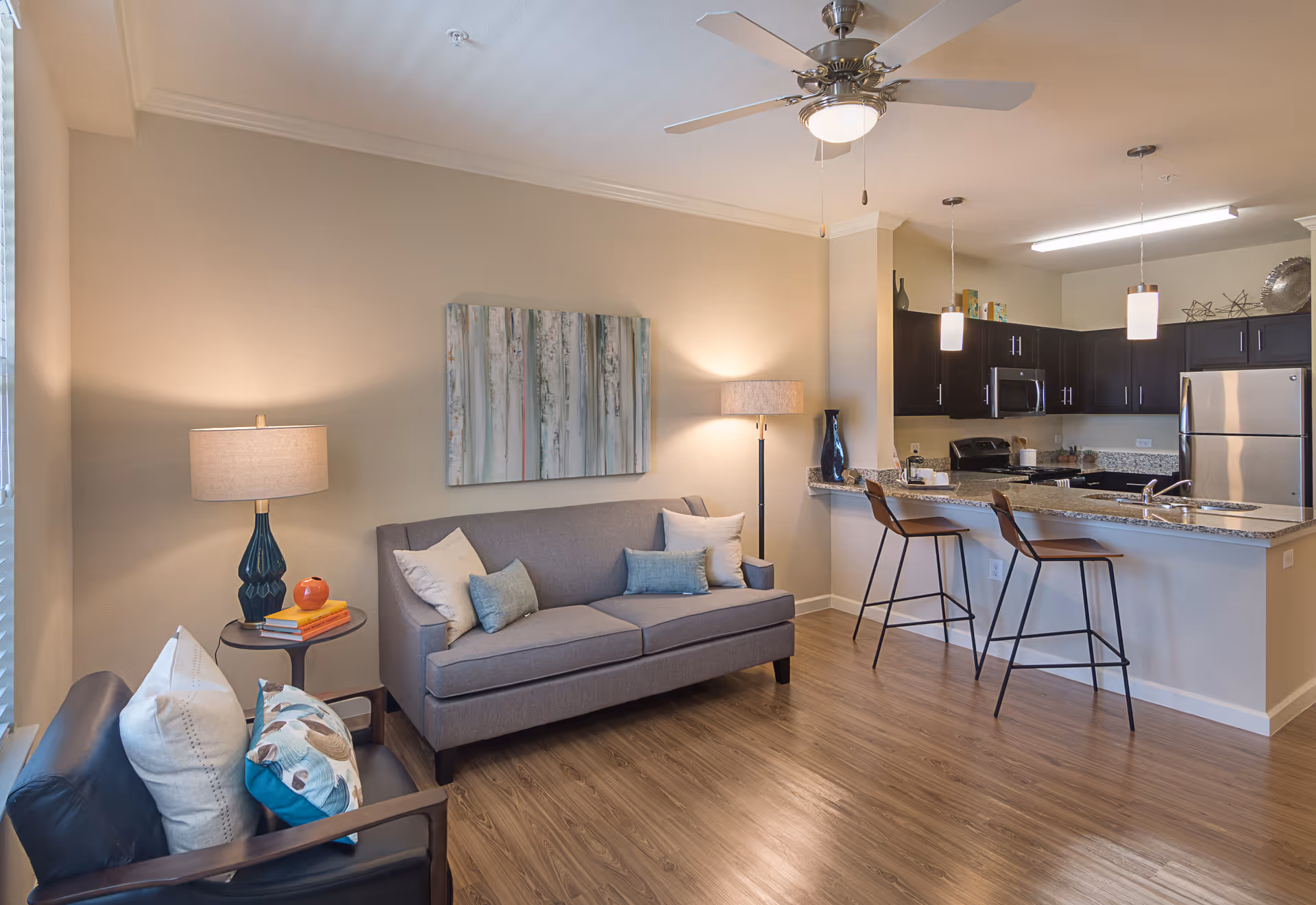 Open-plan living area with a sofa, armchair, lamps, wall art, and a kitchen breakfast bar with two stools.