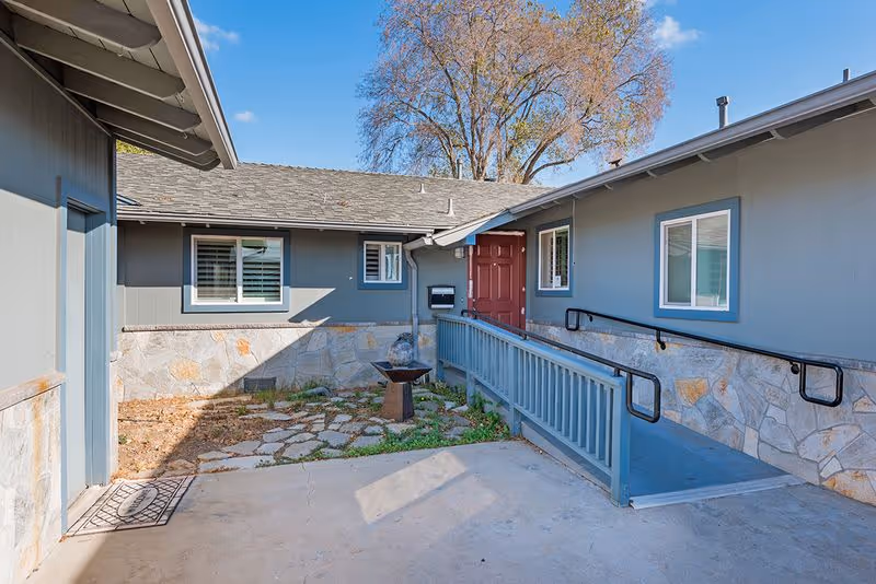 Courtyard entrance to a single-story senior living building with an accessible ramp leading to a red front door.