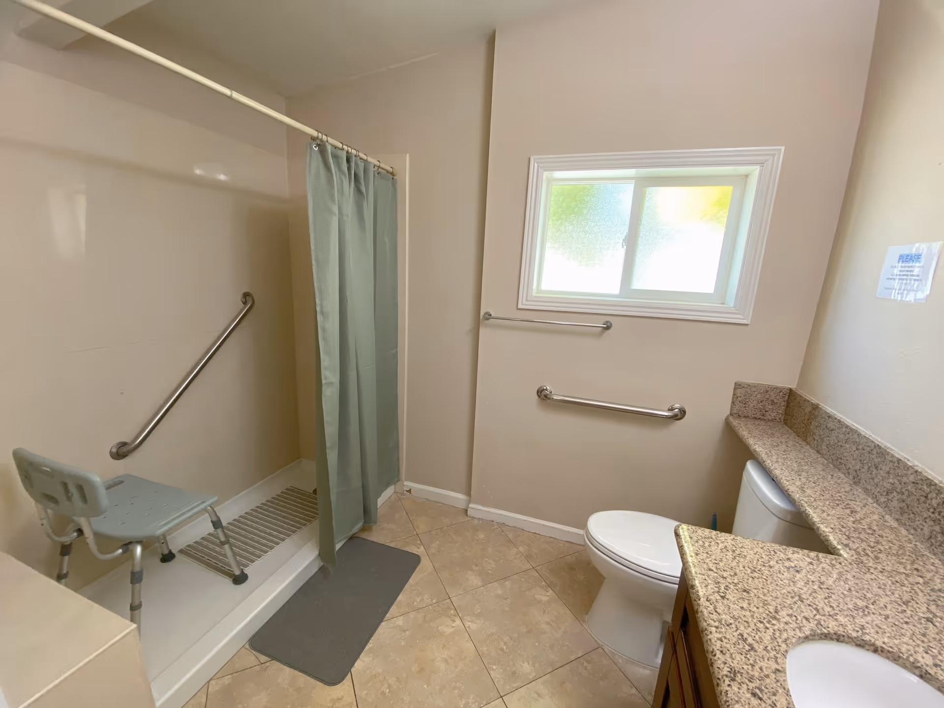 Accessible bathroom with shower, foldable chair, grab bars, toilet, and granite counter.