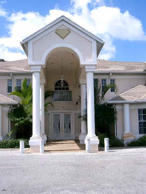 Front exterior view of a two-story building with a covered entrance supported by four white columns. The building is light-colored with a tiled roof, surrounded by greenery including palm trees, under a partly cloudy blue sky.