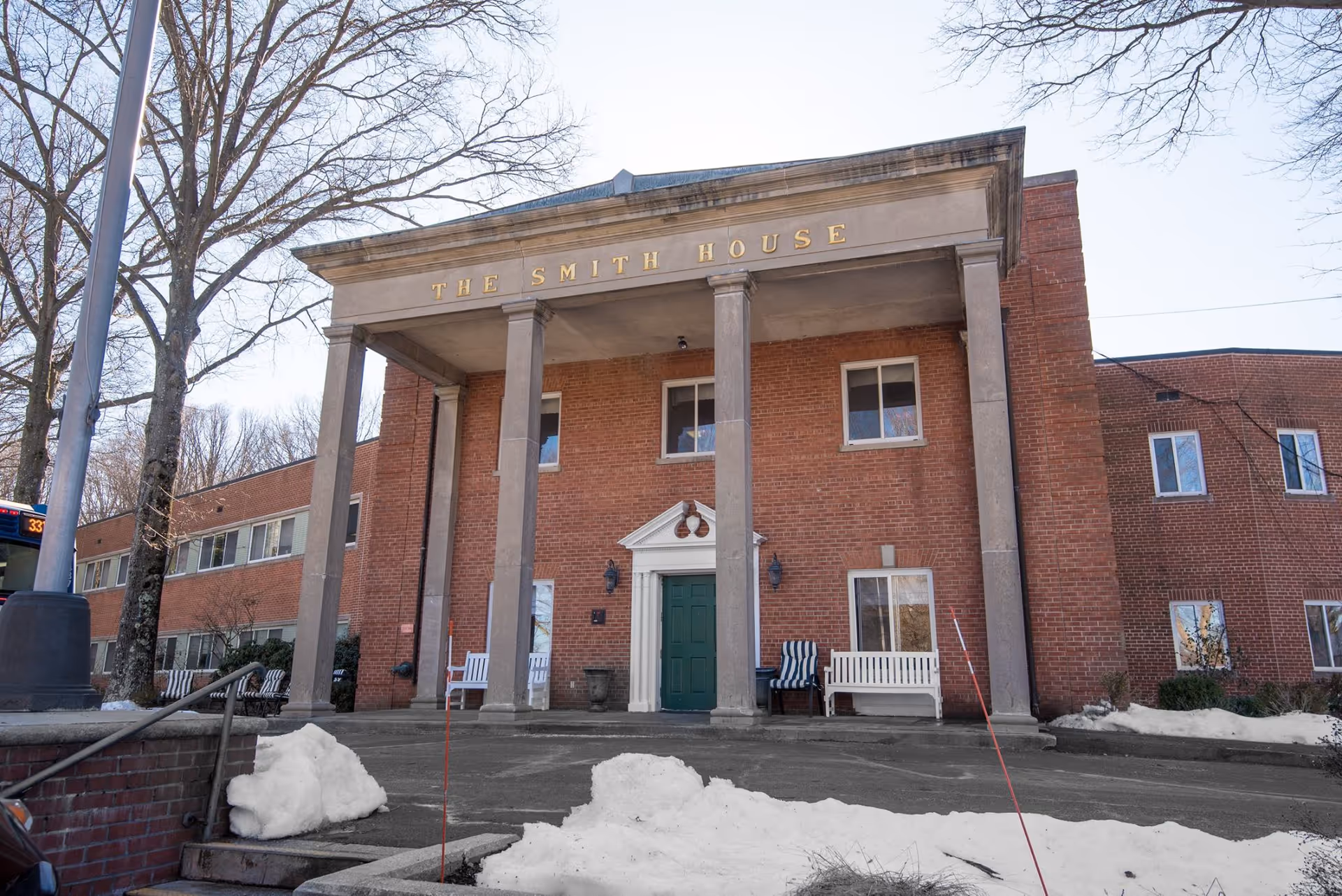 Front facade of a brick building with a columned portico reading 'THE SMITH HOUSE', a green door, benches, and snow piles in front.