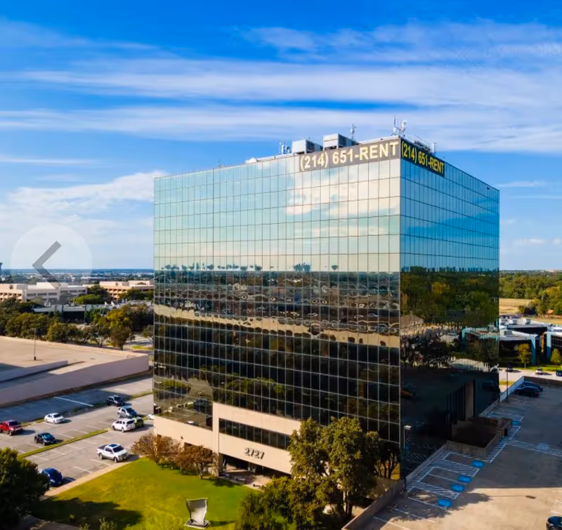 A tall glass office building reflects the surrounding city under a blue sky.
