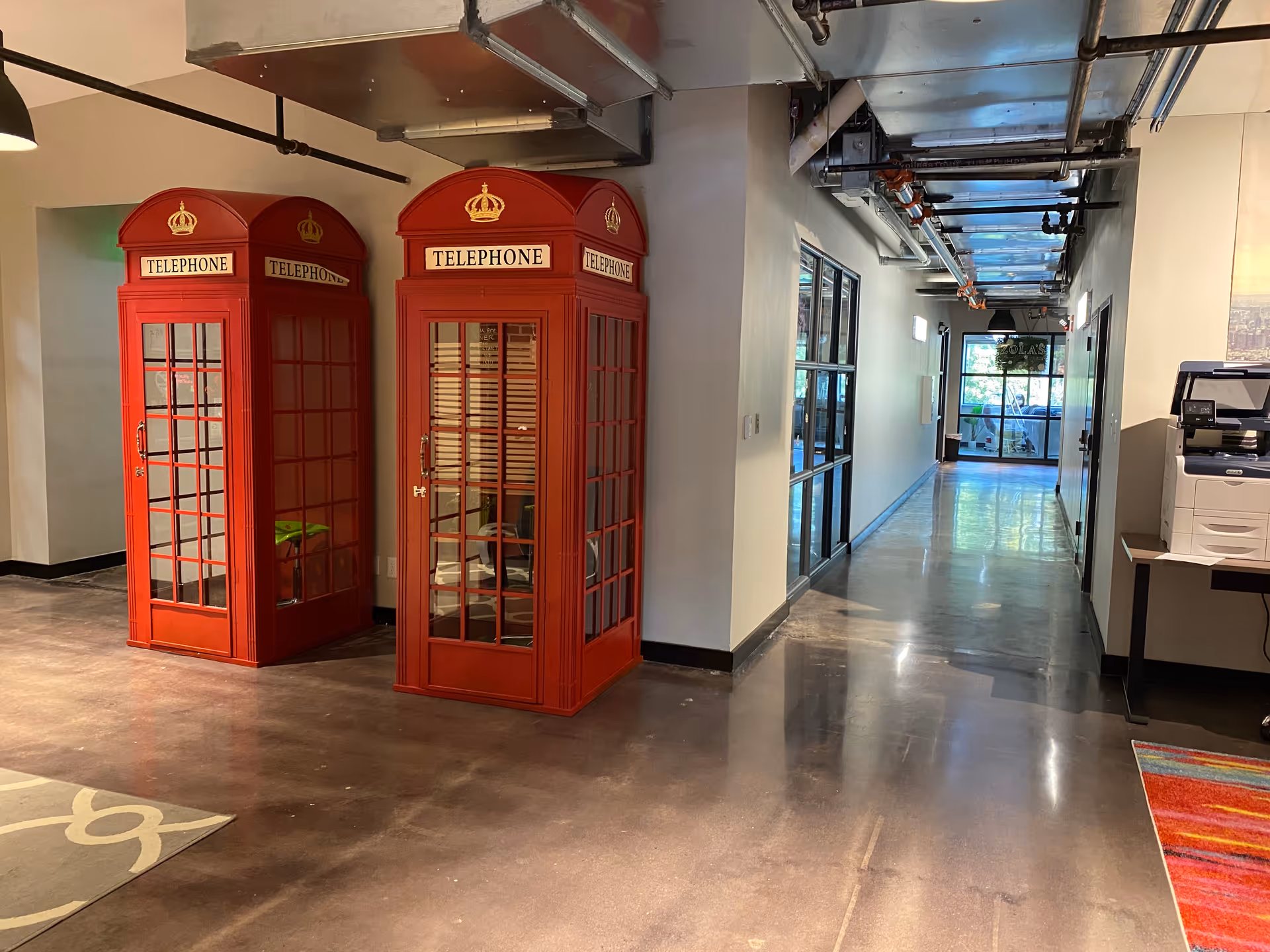 Interior hallway with red vintage telephone booths, exposed pipes, and office equipment.