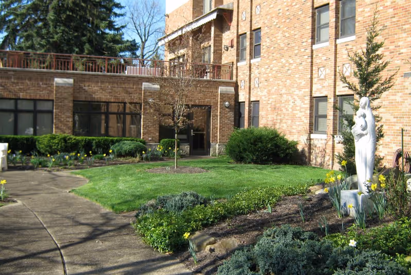 Outdoor garden area at Marian Hall Home featuring a curved concrete pathway, green grass, various shrubs and plants, a small tree, and a white statue of a person holding a child near the brick building.
