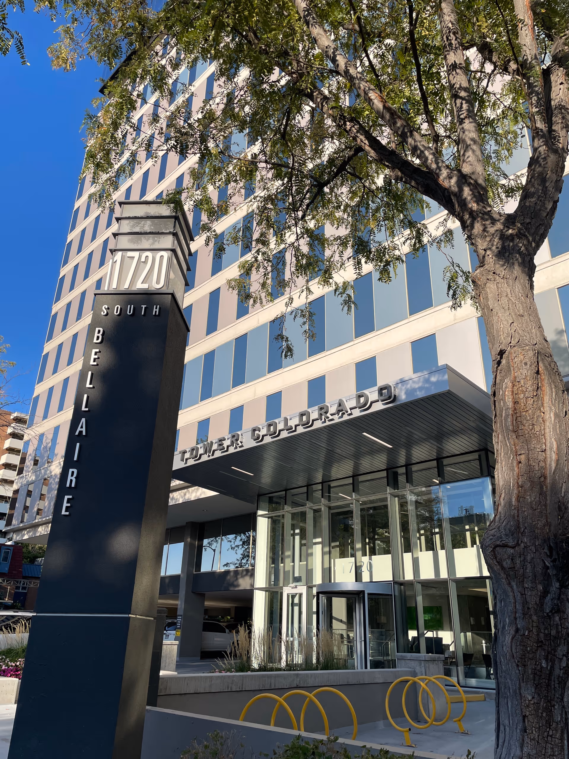 Modern glass-front building entrance with an address pillar and a large tree.
