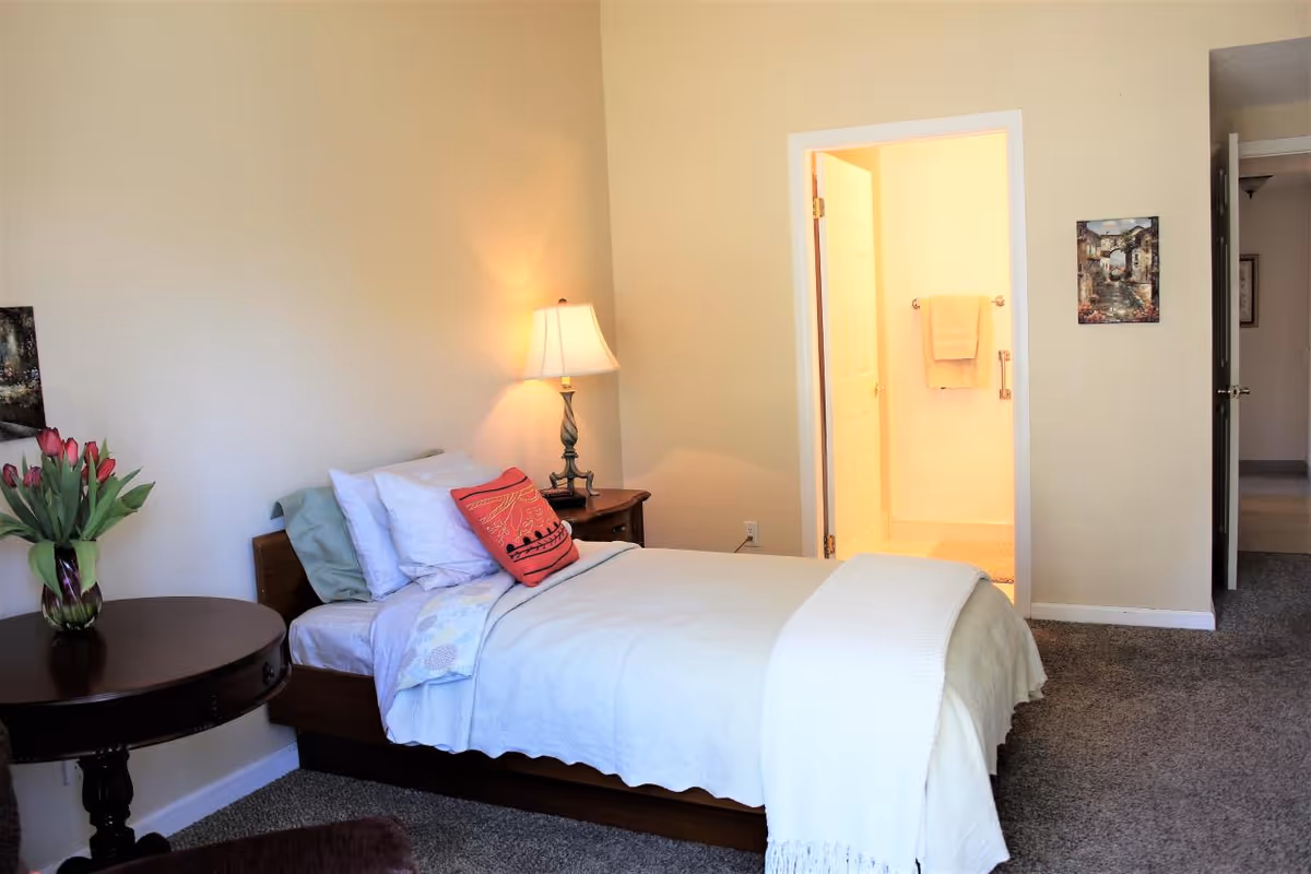 A tidy bedroom in a senior living facility with bed, nightstand, lamp, and flowers.