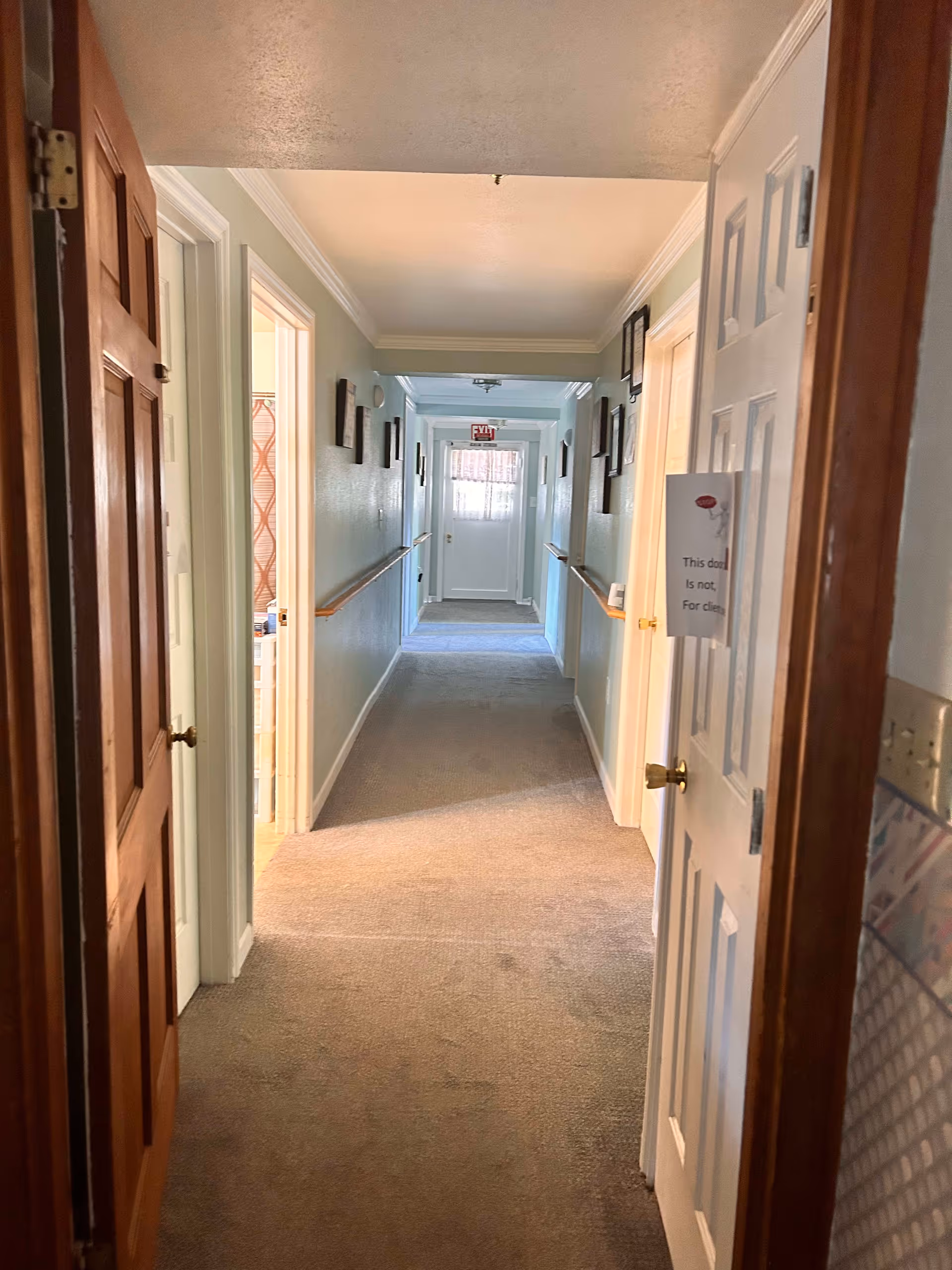 Carpeted interior hallway of a senior home with open doors, wall handrails, framed pictures and an exit door at the far end.