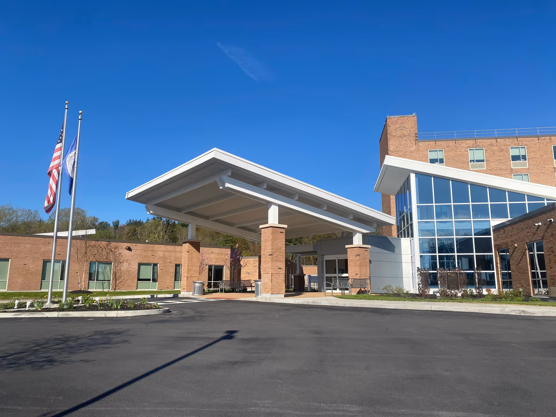 The brick exterior entrance and porte-cochère of a senior living facility with flagpoles and large glass windows under a clear blue sky.