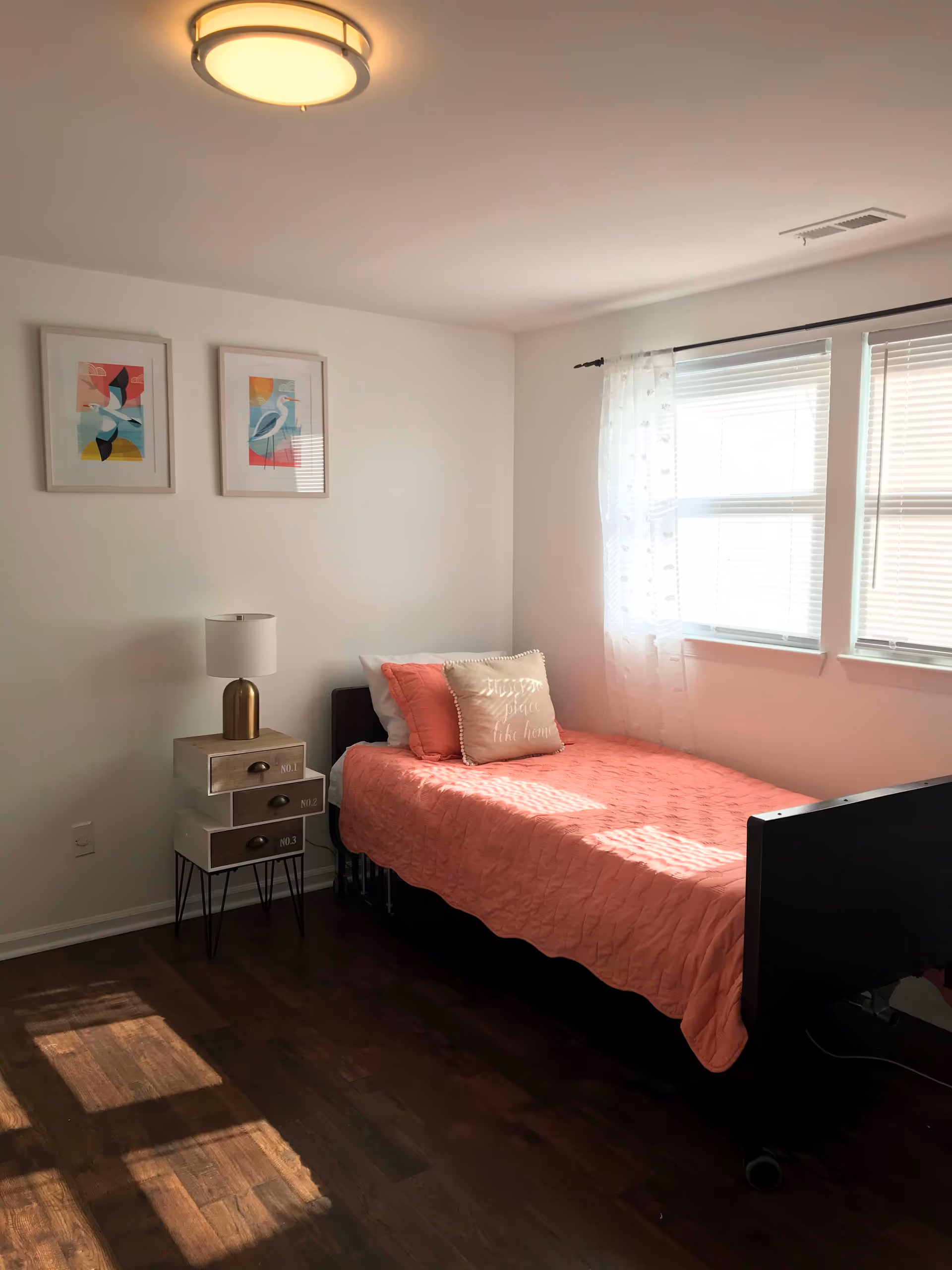A bright bedroom with a single bed covered in a coral quilt and two pillows, one with the text 'my place like home'. Next to the bed is a small nightstand with three drawers labeled No.1, No.2, and No.3, topped with a lamp. Two framed bird artworks hang on the white wall above the nightstand. Sunlight streams through a window with white blinds and sheer curtains, casting shadows on the dark wooden floor.