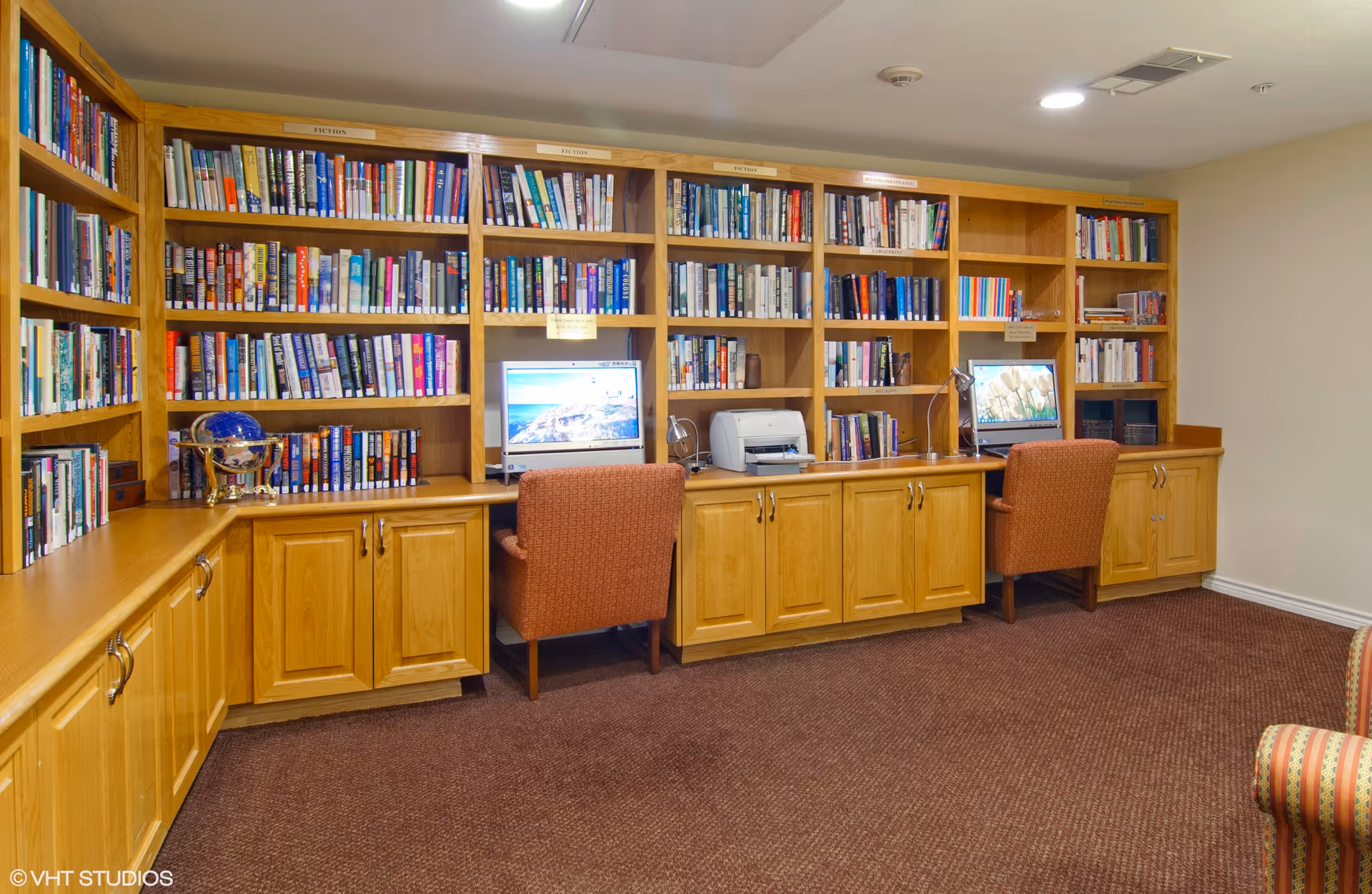A library-style room with wooden bookshelves, two computer workstations and upholstered chairs.