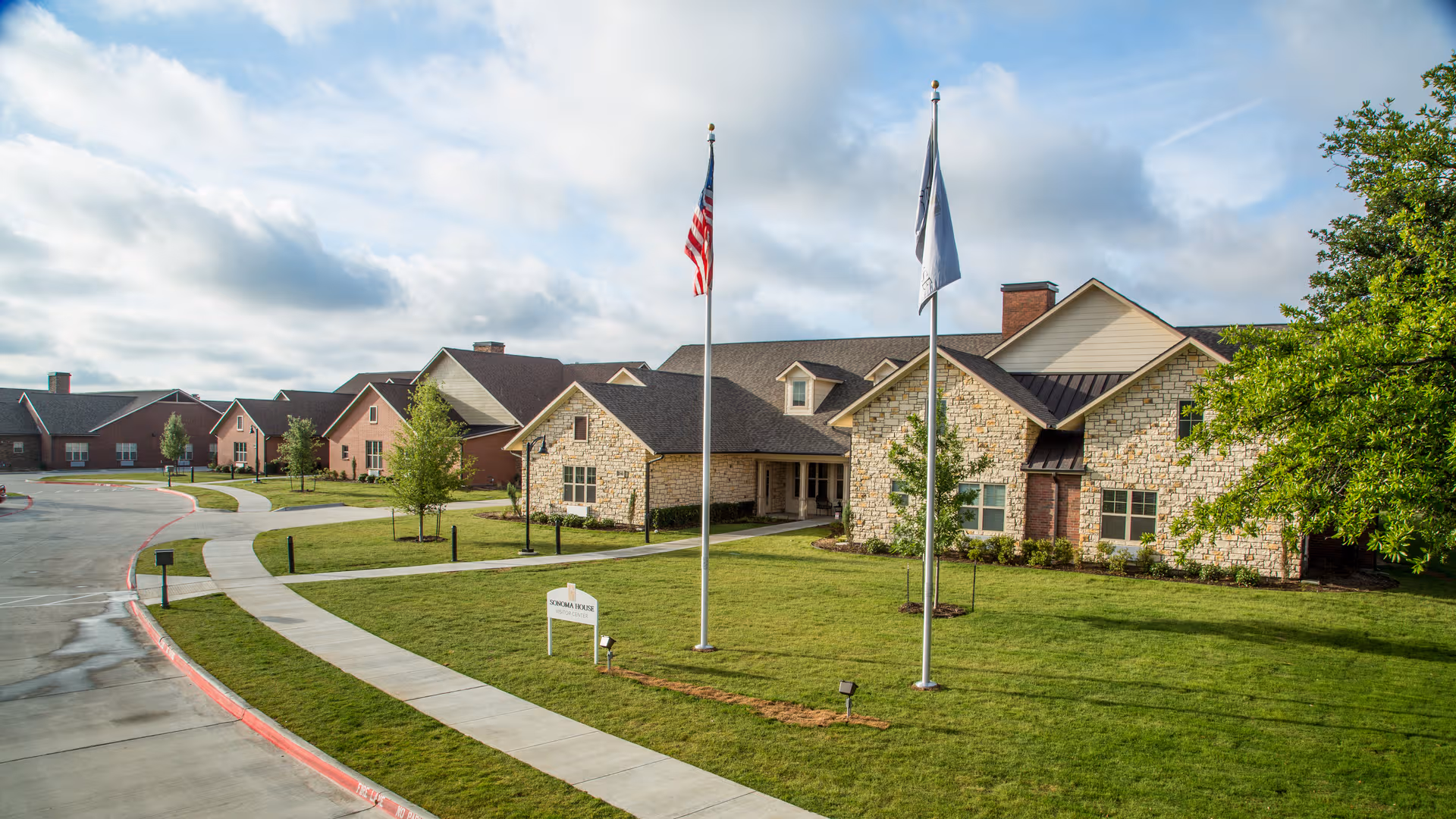 Exterior view of Sonoma House Assisted Living with flagpoles, a curved walkway, and a green lawn.