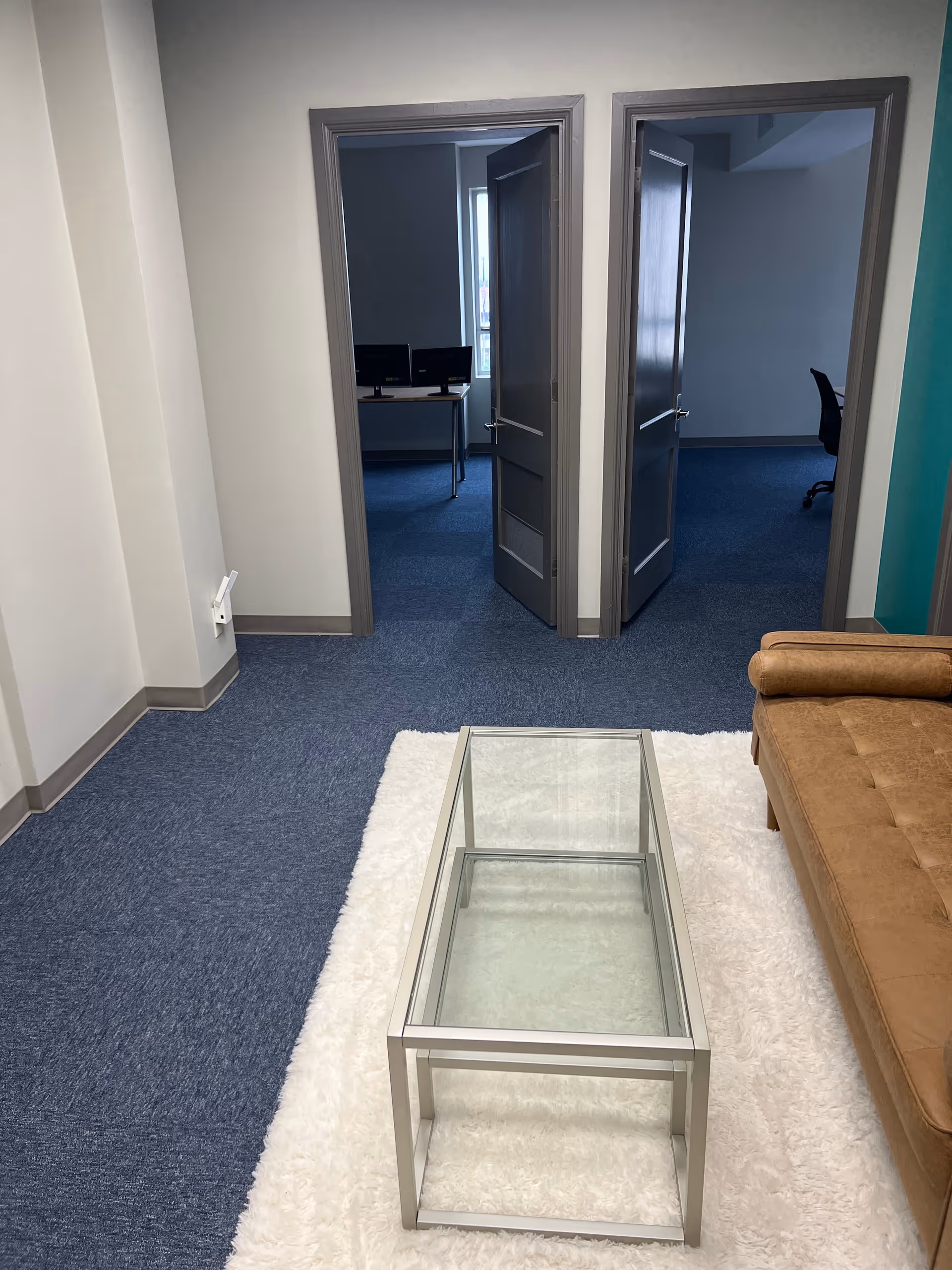 Interior lounge area with glass coffee table, white rug, blue carpet, and desks visible beyond doors.