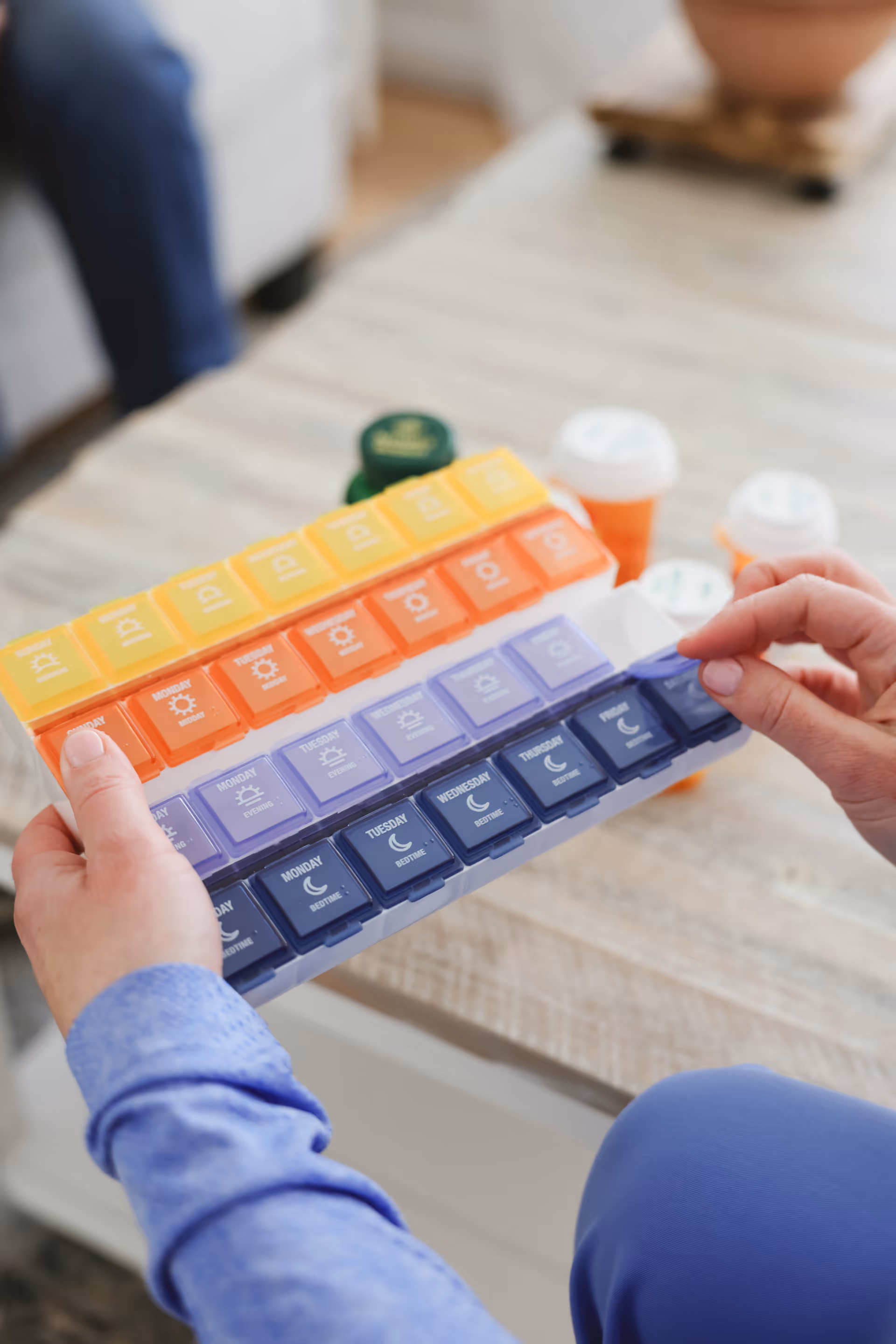 A person’s hands hold a colorful, multi-day pill organizer on a wooden table indoors.